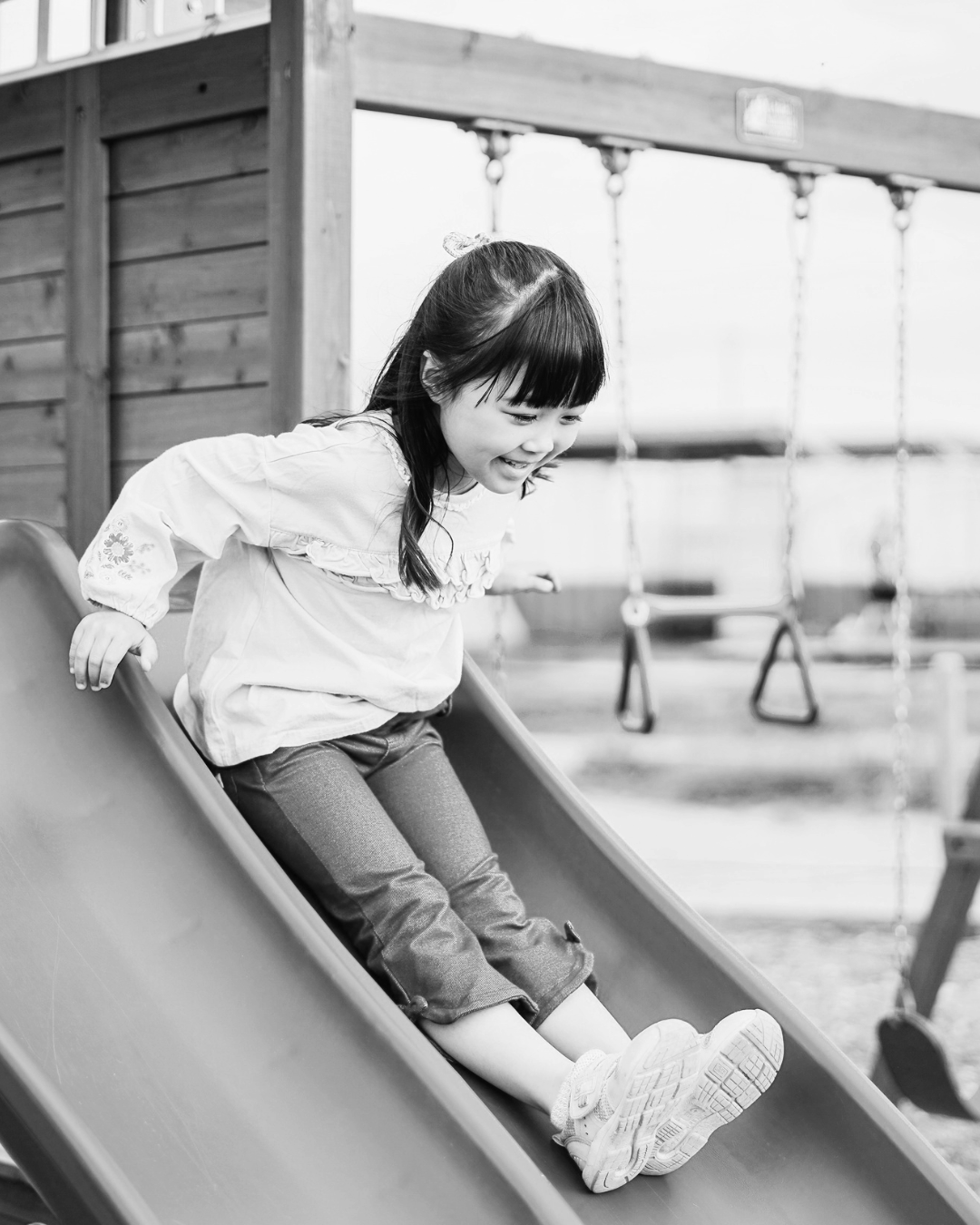 A young girl with dark hair, smiling and playing on a slide at an outdoor playground.