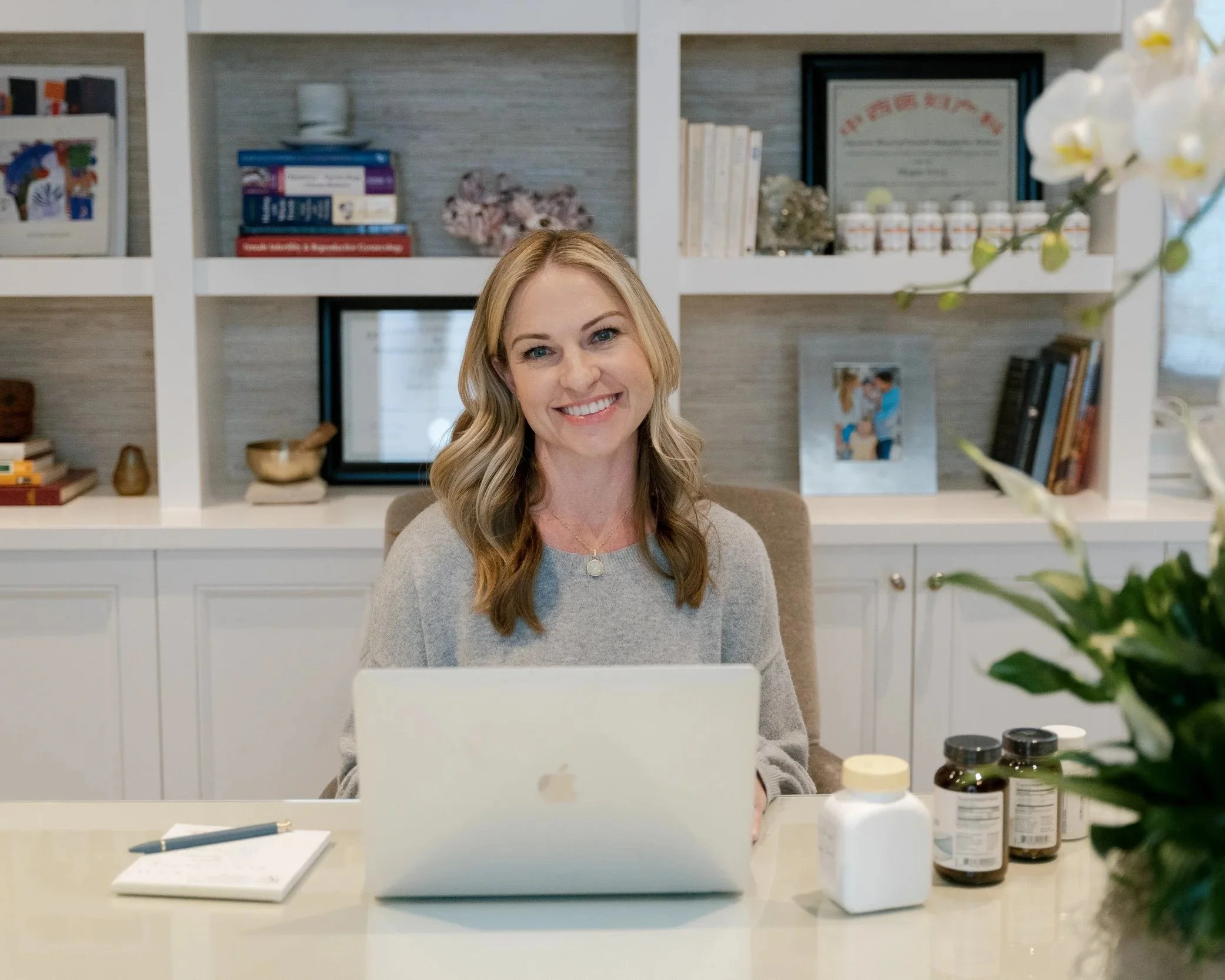 Megan Joyce, LAc FABORM sitting at a desk in her office with a laptop, notebook, and pen in front of her, in a room with white shelves filled with books, framed degree certificates, decorative items, and a window in the background.