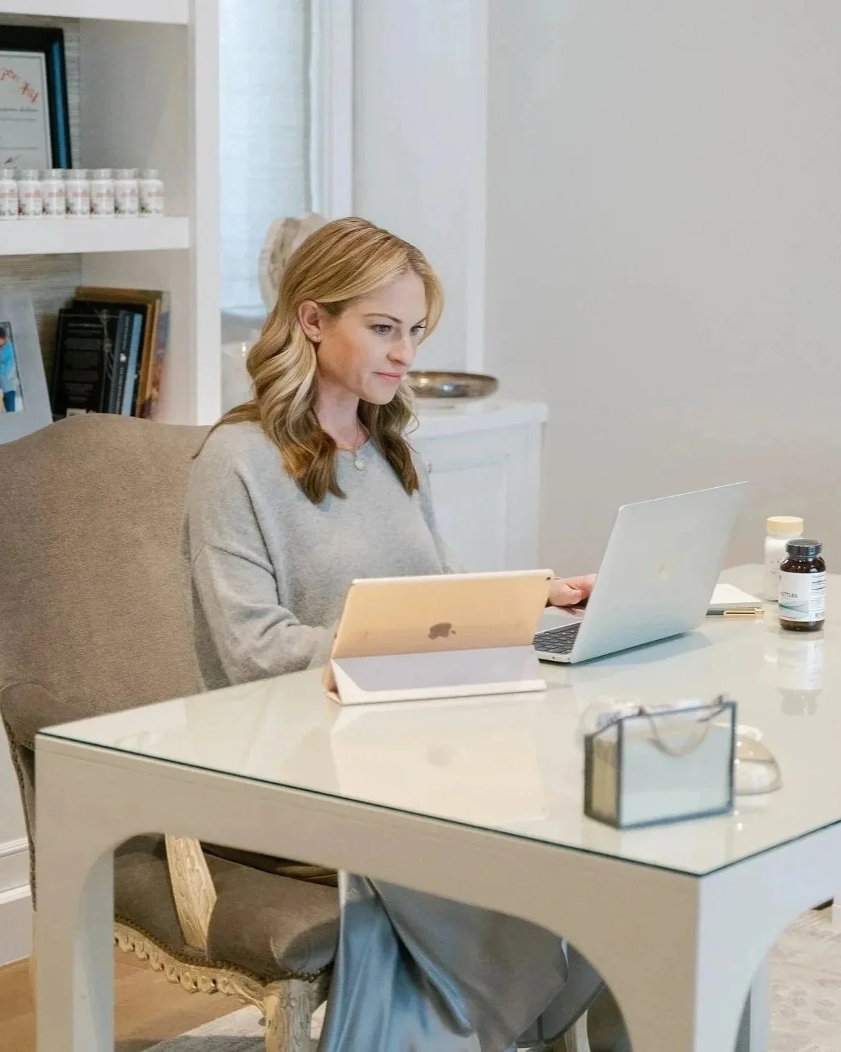 Woman with blonde hair working at a desk using a laptop and a tablet in a home office.