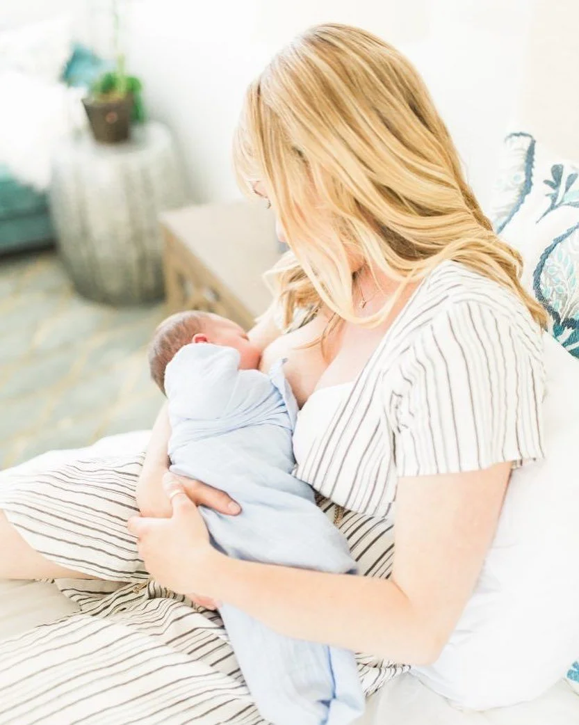 A woman (Megan Joyce) with blonde hair is breastfeeding a newborn baby in a cozy, well-lit room, sitting on a bed with patterned pillows.