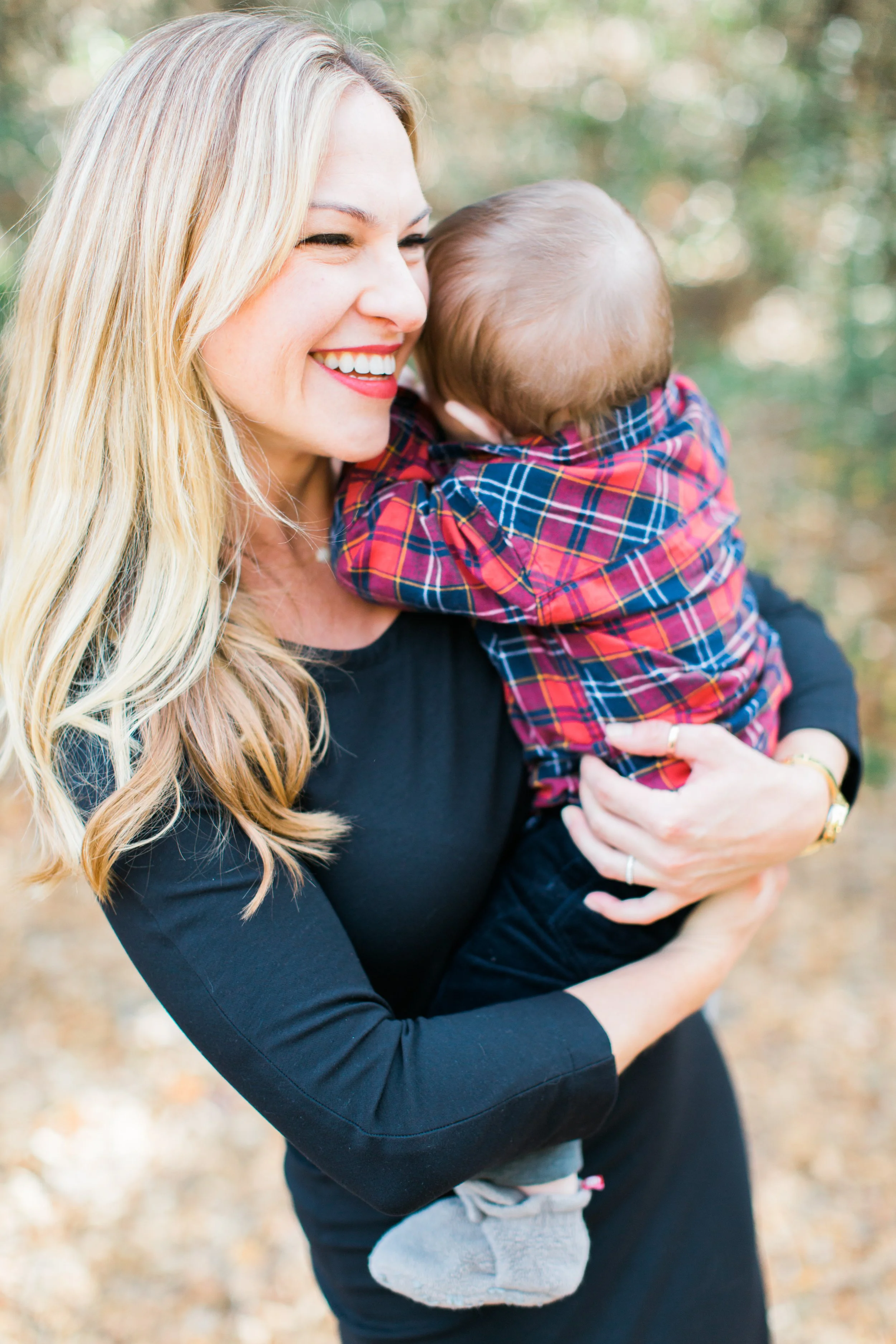 Megan Joyce, LAc holding a young baby boy with light brown hair, dressed in a red and blue plaid shirt. The background shows a blurred outdoor setting with trees and fallen leaves.