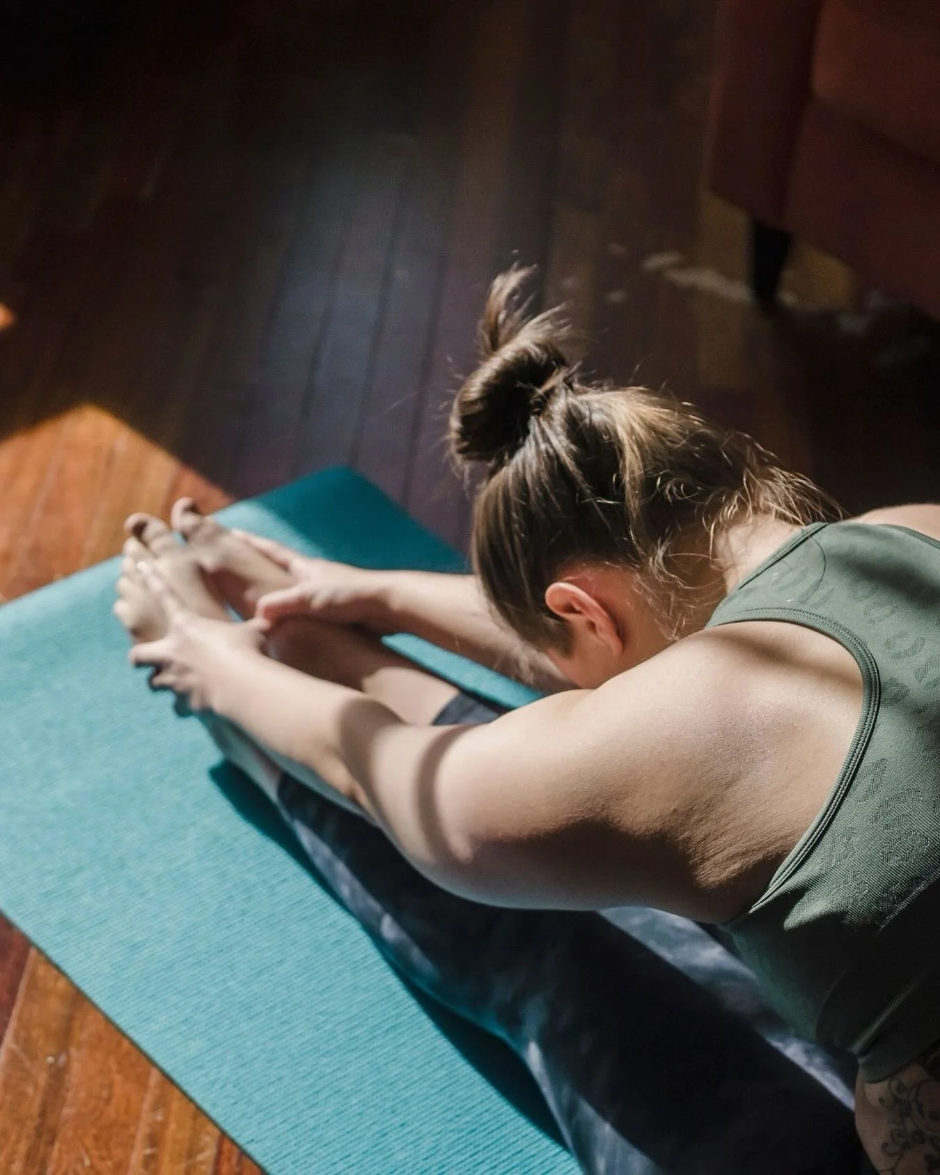 A woman with brown hair tied in a bun practicing yoga, stretching forward on a teal yoga mat on a wooden floor.