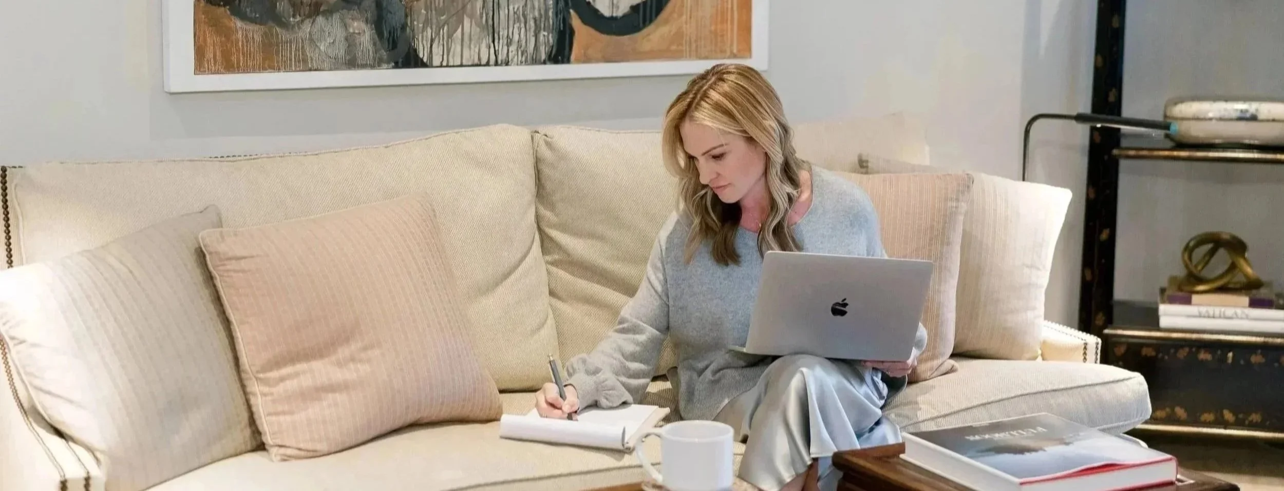 A woman (Megan Joyce) sitting on a beige couch with pink cushions, working with a laptop and writing notes in a notebook. There is a coffee mug and a book on the table in front of her, and an art piece on the wall behind her.