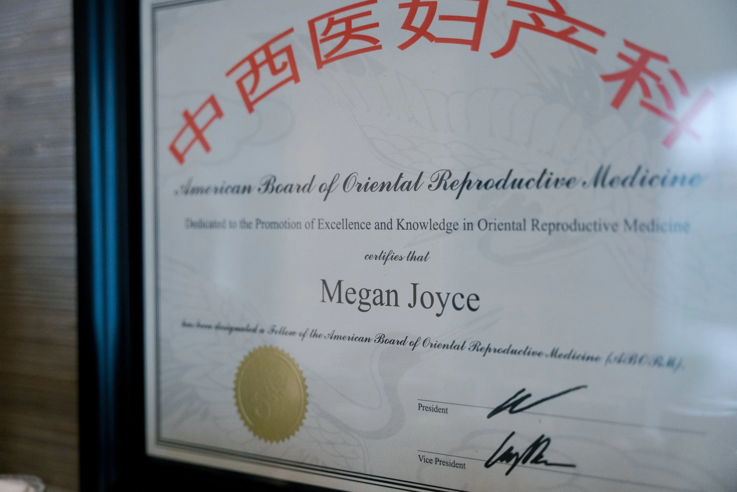 Close-up of a certificate from the American Board of Oriental Reproductive Medicine with red Chinese characters at the top. The certificate awards Megan Joyce, with black text, and includes a gold seal at the bottom left. It is signed by the President and Vice President.