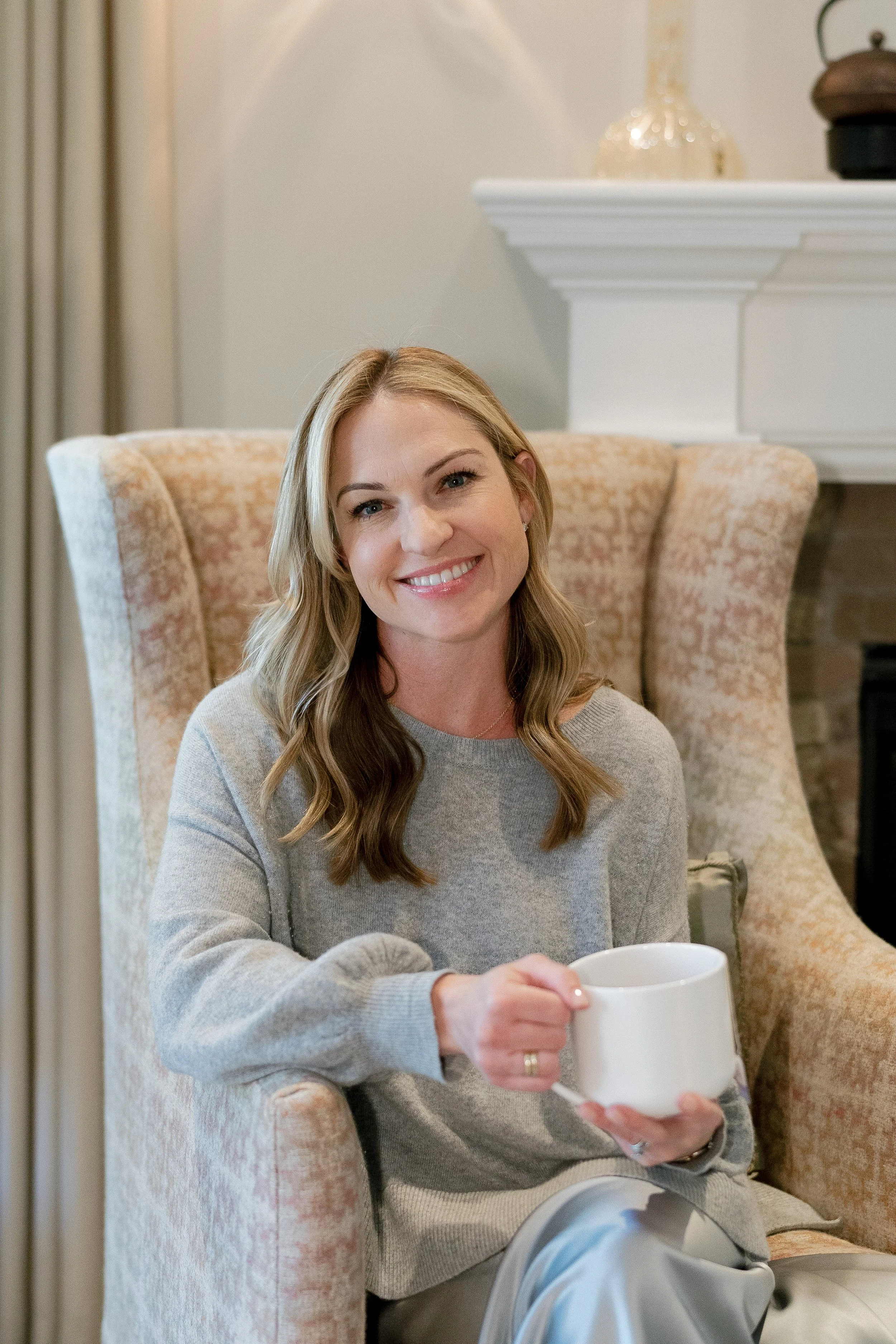 Megan Joyce smiling and sitting in an armchair, holding a white coffee mug, in a cozy living room with a fireplace in the background.