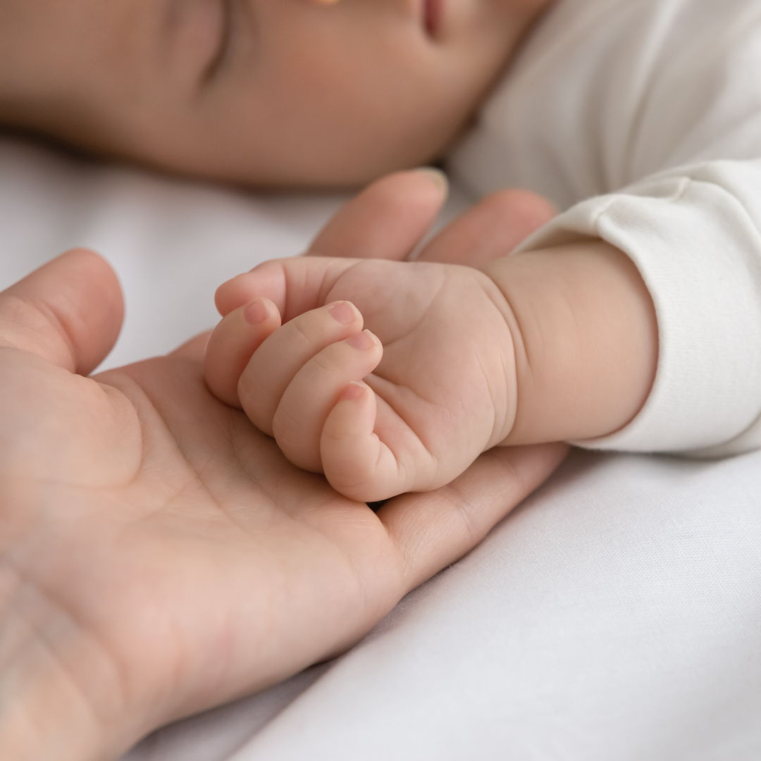 A baby's hand holding an adult's hand, with the baby's face partially visible in the background.