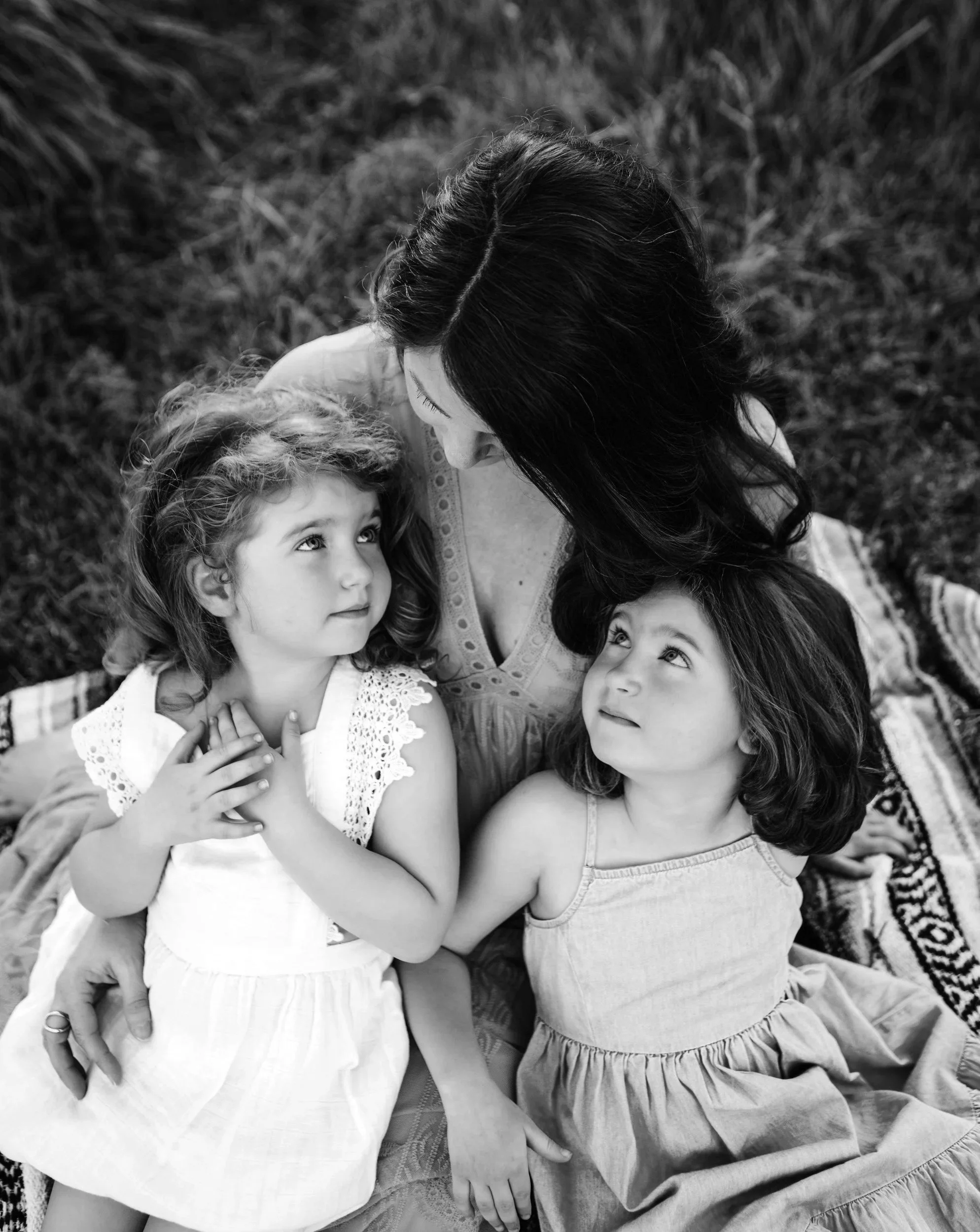Black and white photo of a woman with three children, sitting on grass outside. The woman is looking down at the children, who are looking up at her. The children are a girl with curly hair and a girl with straight hair. The woman has long dark hair, and one girl has long wavy hair, another has shorter curly hair, and the third girl has straight hair with bangs.