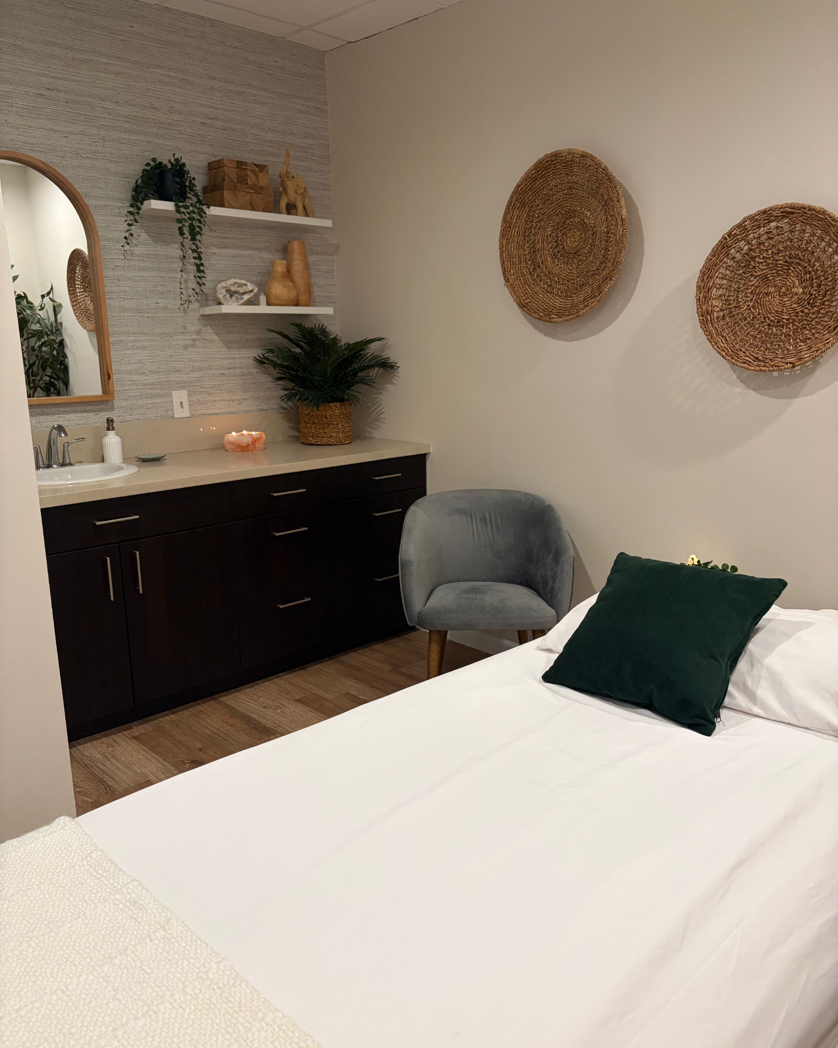 Cozy bedroom with a white bed, dark green pillow, gray accent chair, dark wood dresser, woven wall baskets, mirror, plants, and decorative shelves