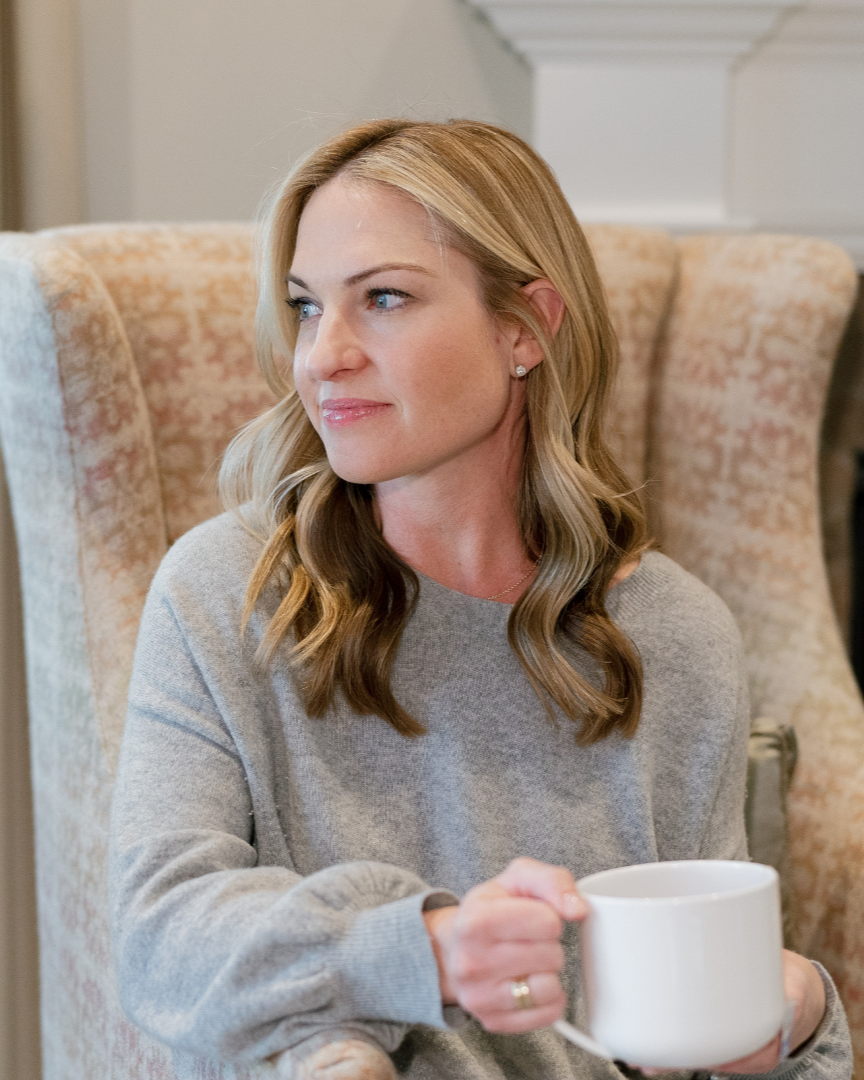 Megan Joyce sitting in an armchair, holding a white mug, wearing a gray sweater, looking to her left.