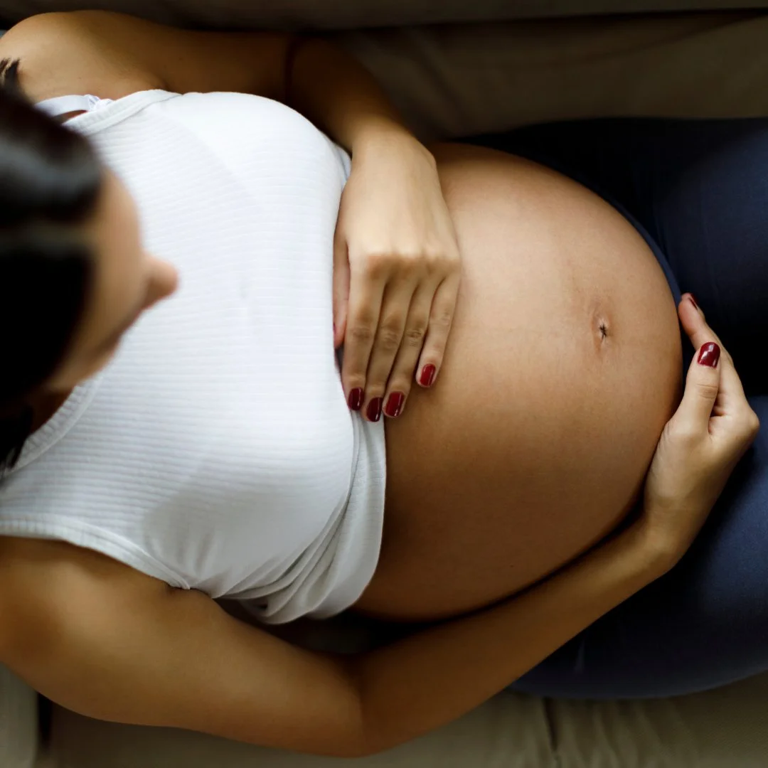 A pregnant woman lying down on a couch, touching her bare belly with one hand, wearing a white sleeveless top and dark pants.
