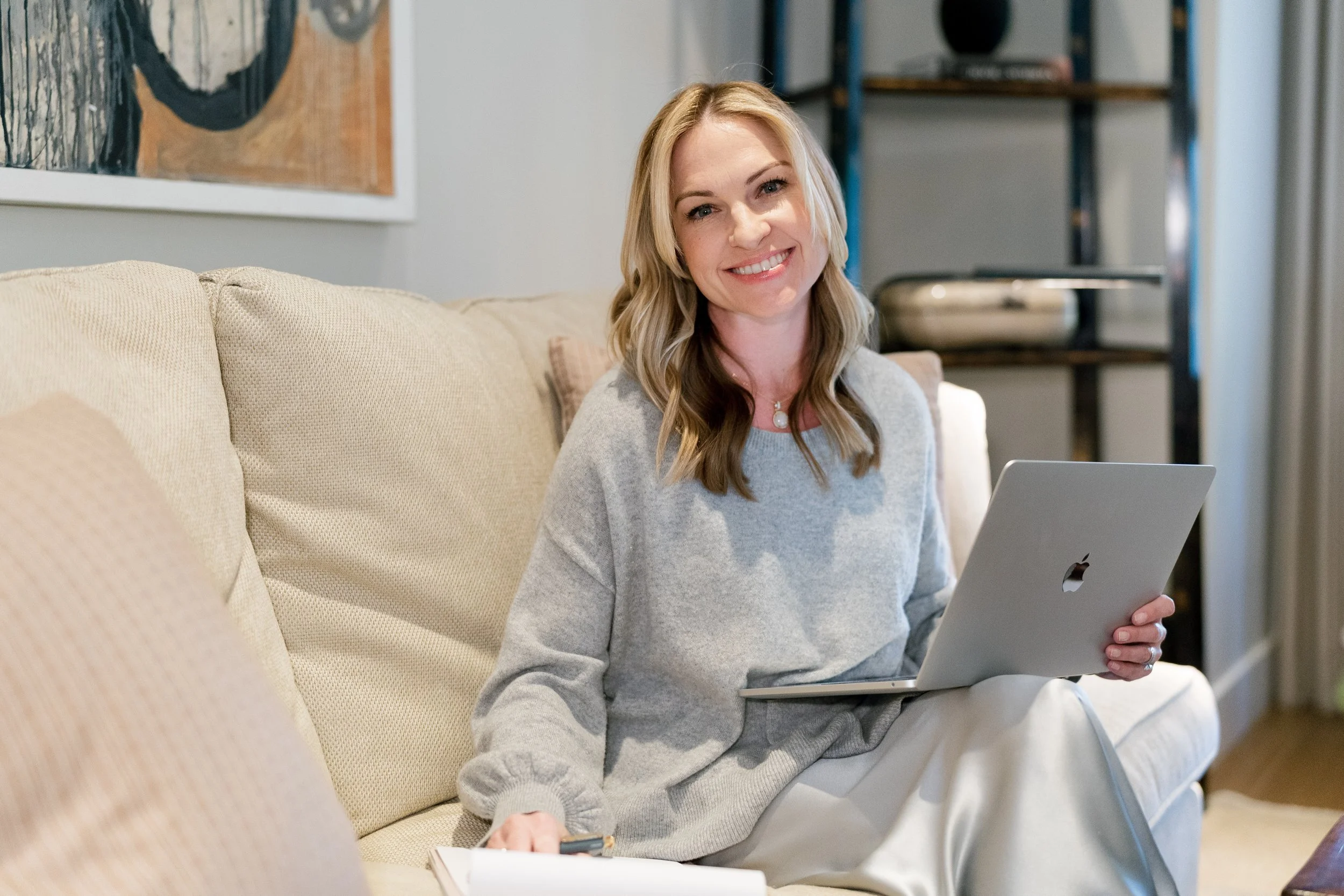 A smiling woman (Megan Joyce) with wavy blonde hair sitting on a beige couch, holding a silver laptop and looking at the camera in a cozy living room.