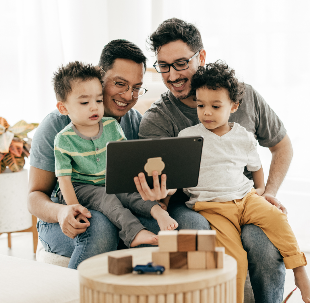 Two adults and two young children sitting together on a sofa, looking at a tablet device, in a bright living room with wooden blocks on a table in front.