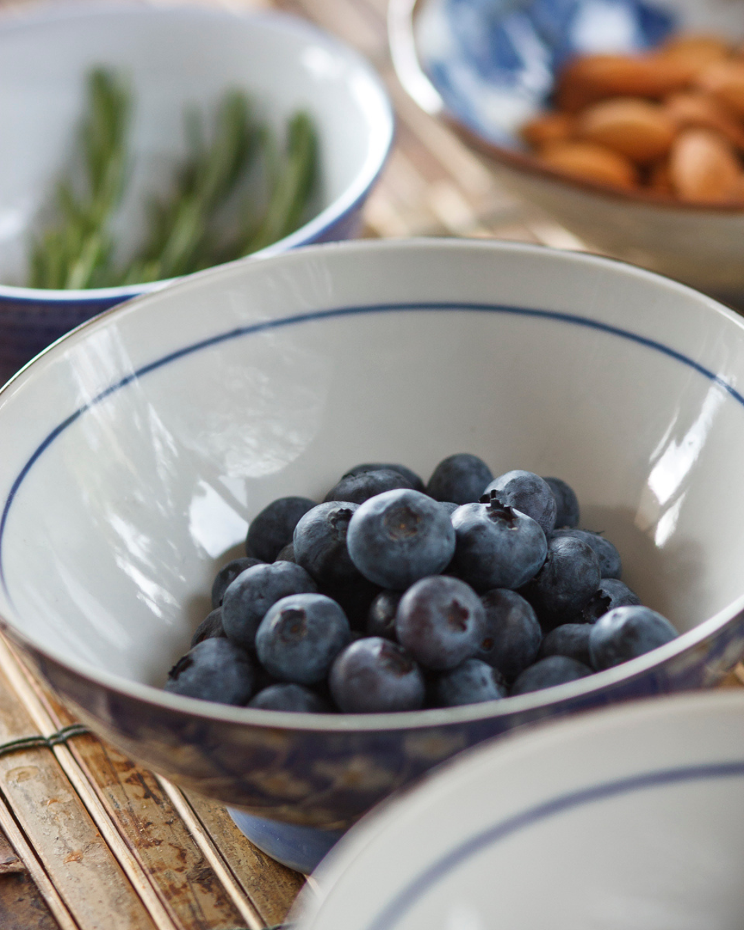 A bowl of fresh blueberries on a wooden surface with other bowls of different ingredients in the background.