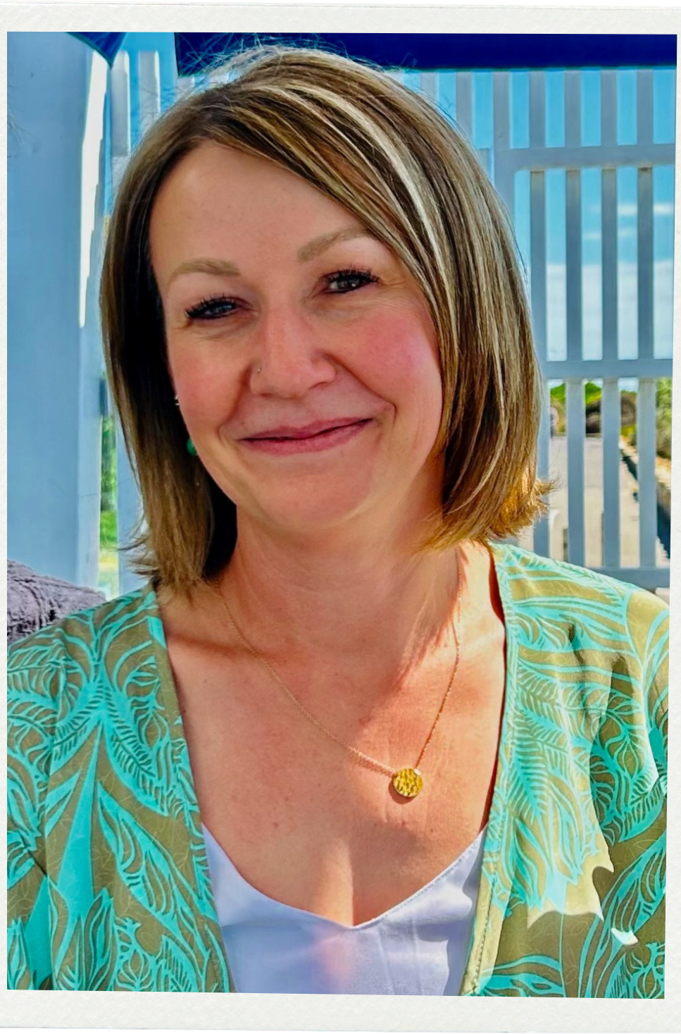 A smiling woman with short brown hair and wearing a blue and green leafy patterned top, a white shirt, and a gold necklace, standing in front of a blue fence and greenery.