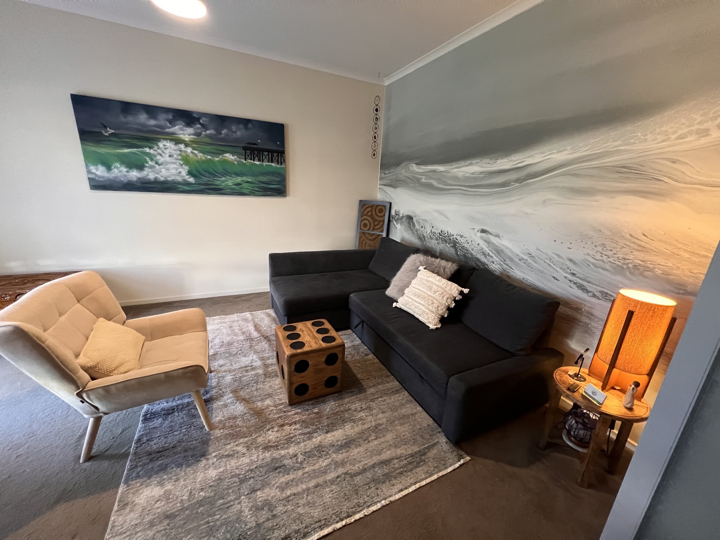 Living room with a beige armchair, black sofa with cream and gray pillows, wooden cube table, artwork of an ocean wave and pier on the wall, corner lamp with orange shade, and a large beige and gray area rug.