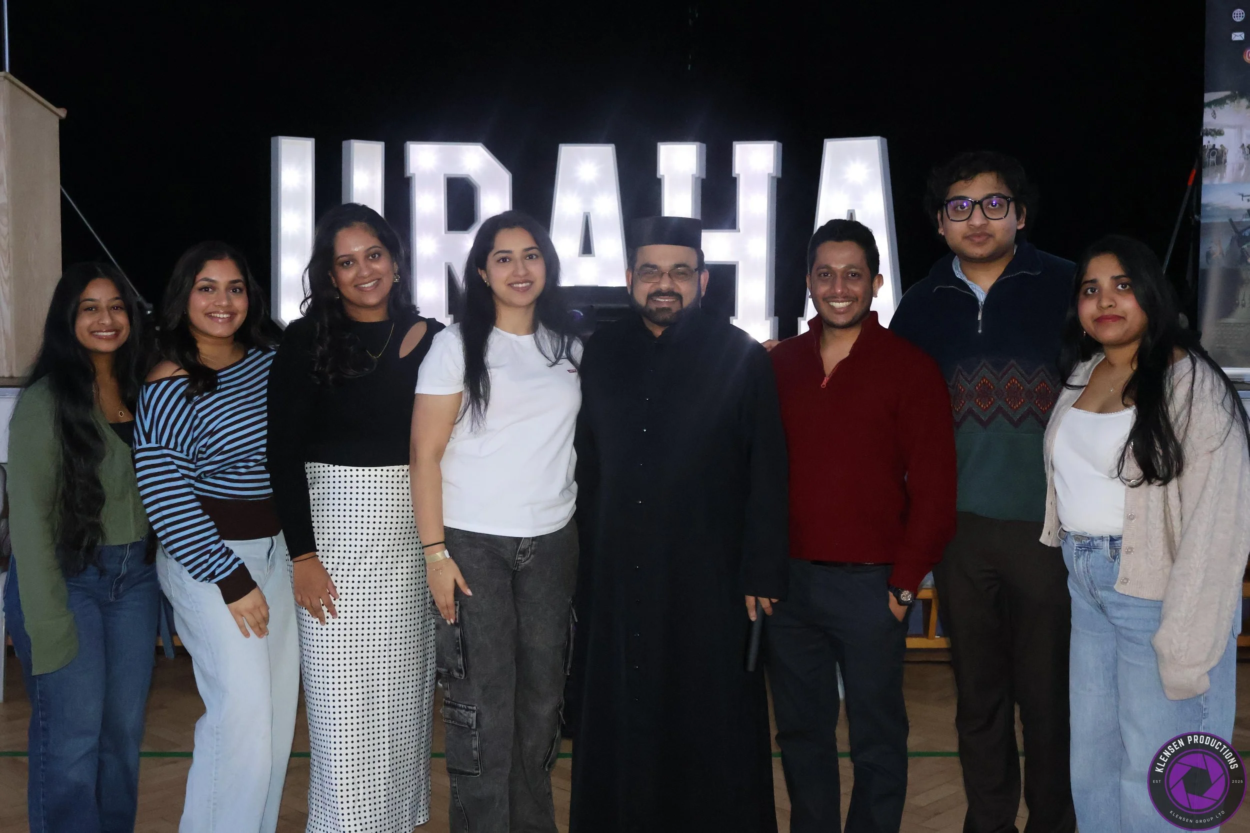 Group of nine people standing together indoors, smiling, with large illuminated letters spelling 'URAAHA' in the background.