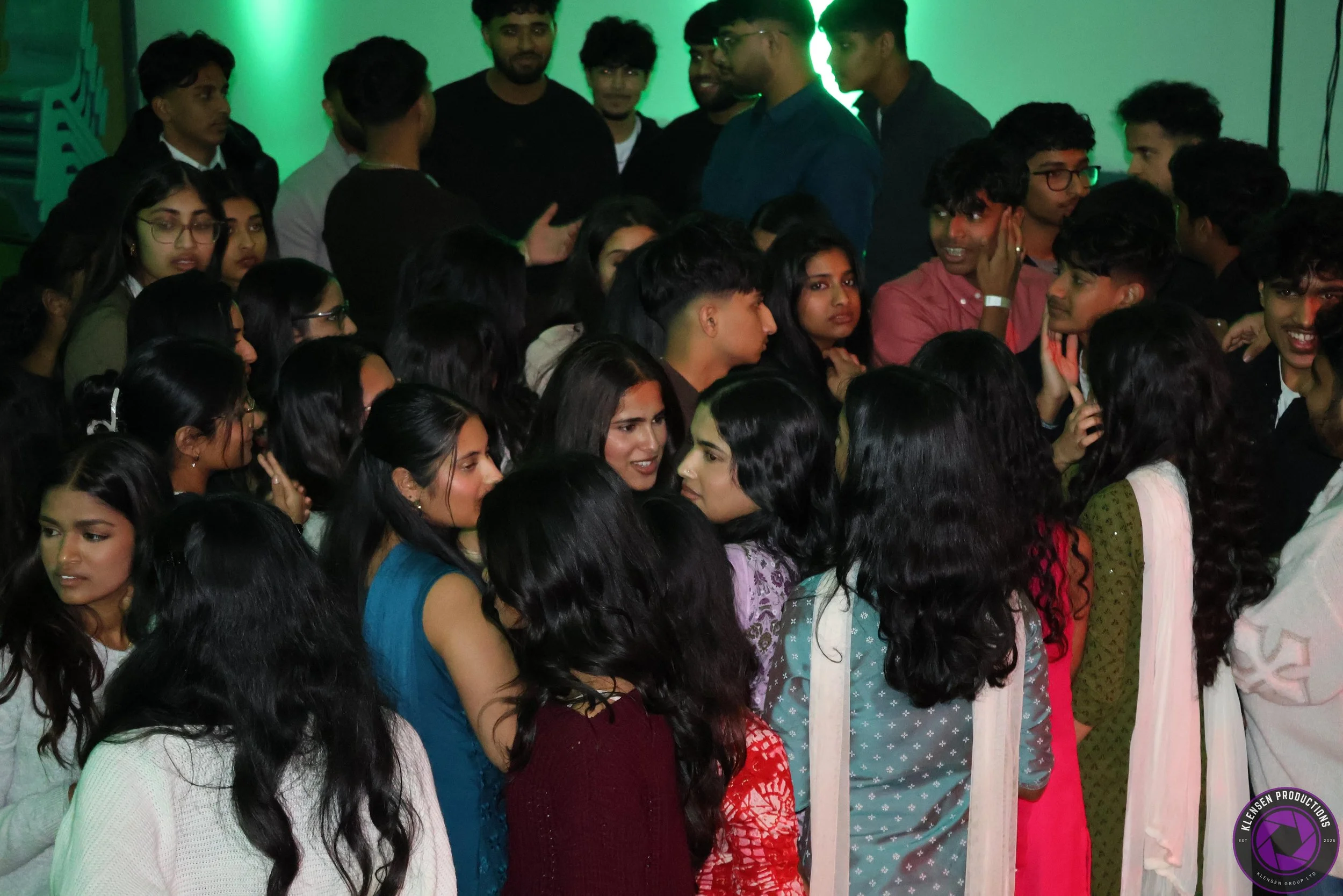A group of young people gathered indoors, some engaged in conversation, with green lighting in the background.