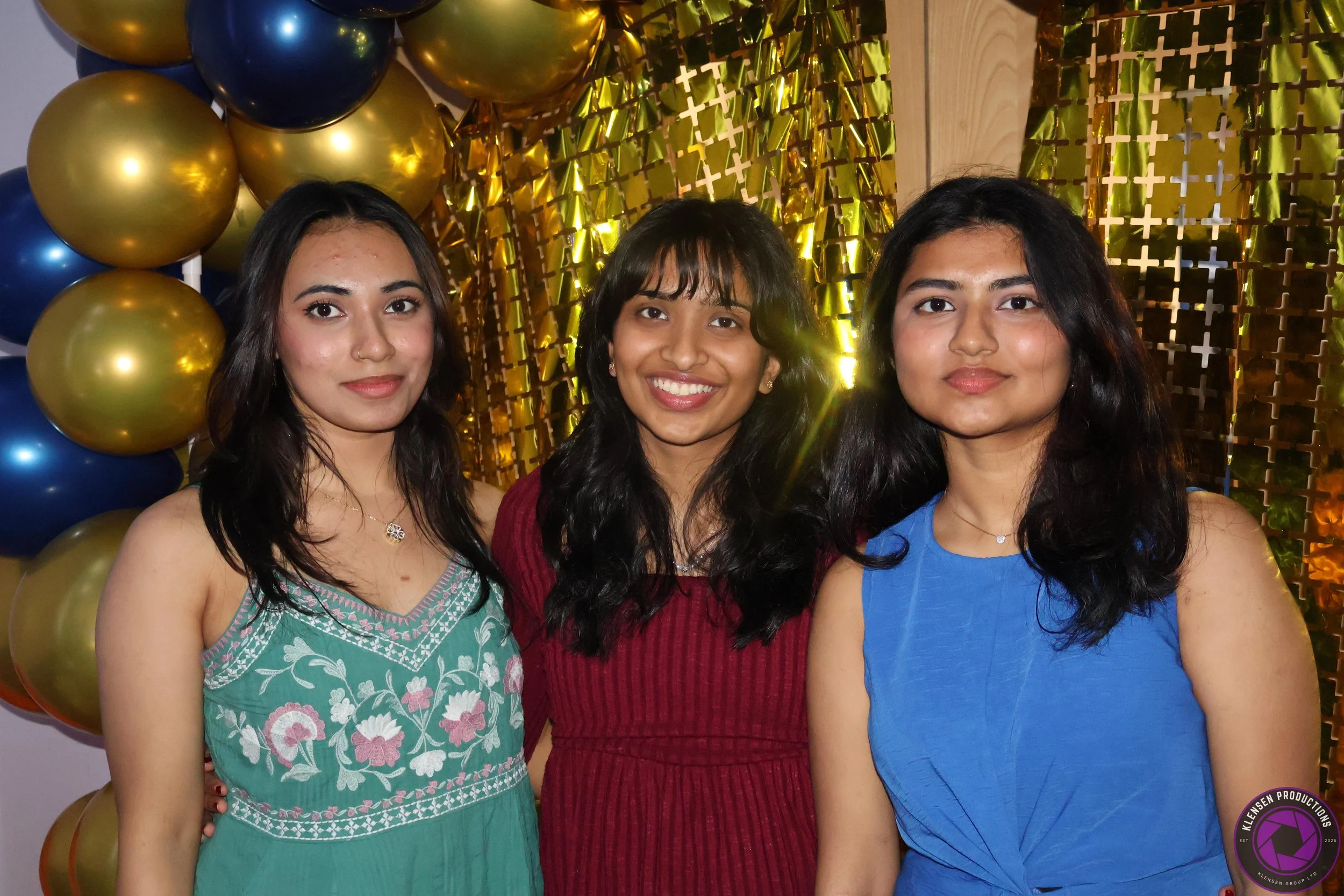 Three young women standing close together at a celebration, smiling, with gold and blue balloons and a golden backdrop behind them.