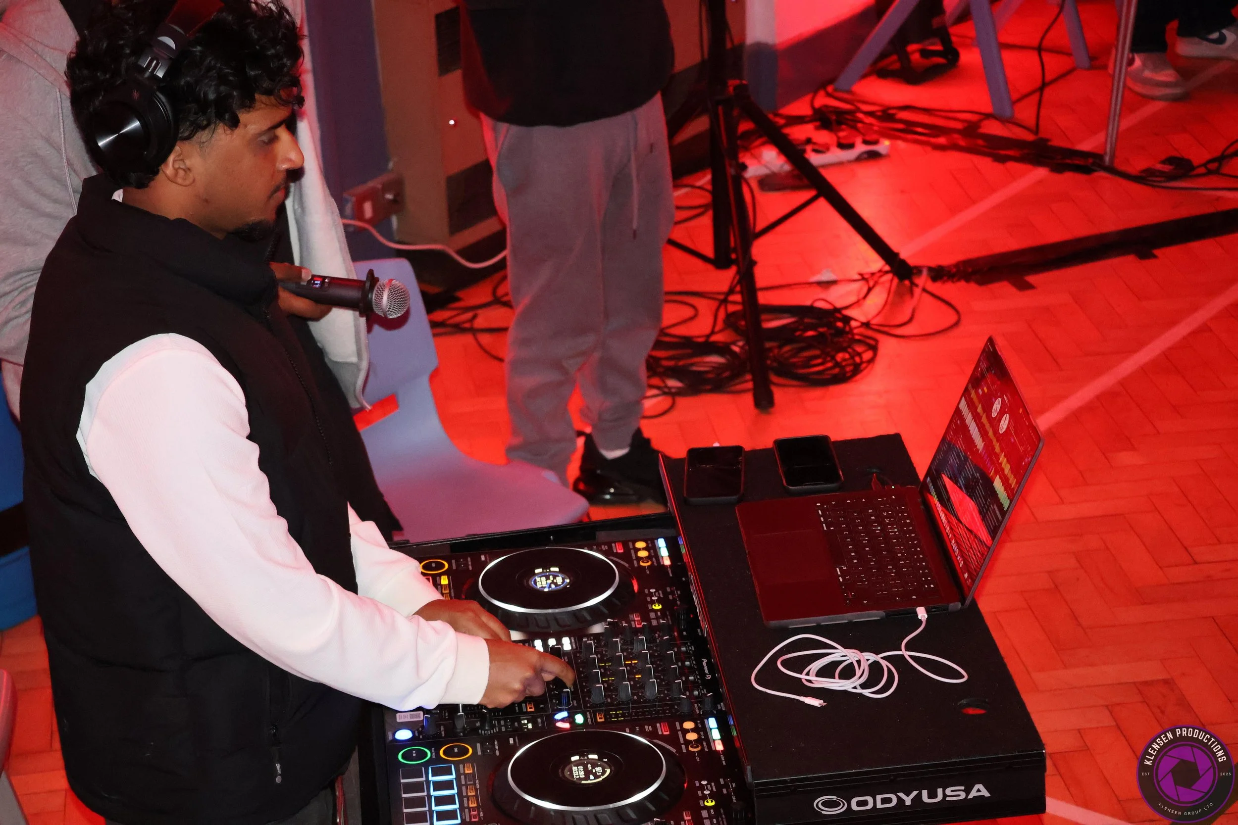 A DJ wearing headphones and a black vest over a white shirt is using a DJ controller with colorful lights. There is a laptop, two smartphones, and earphones on the table. The background has red lighting, cables, and a wooden floor.