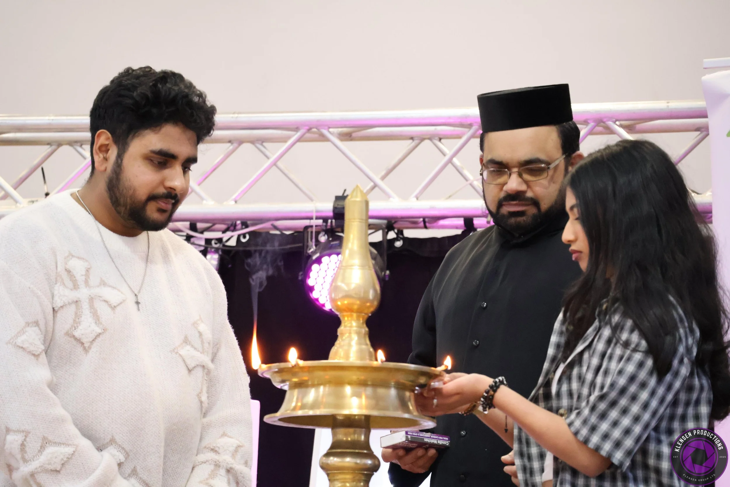 Three people participating in a traditional lighting ceremony involving a brass lamp with flames, with a backdrop of pink and white stage lighting.
