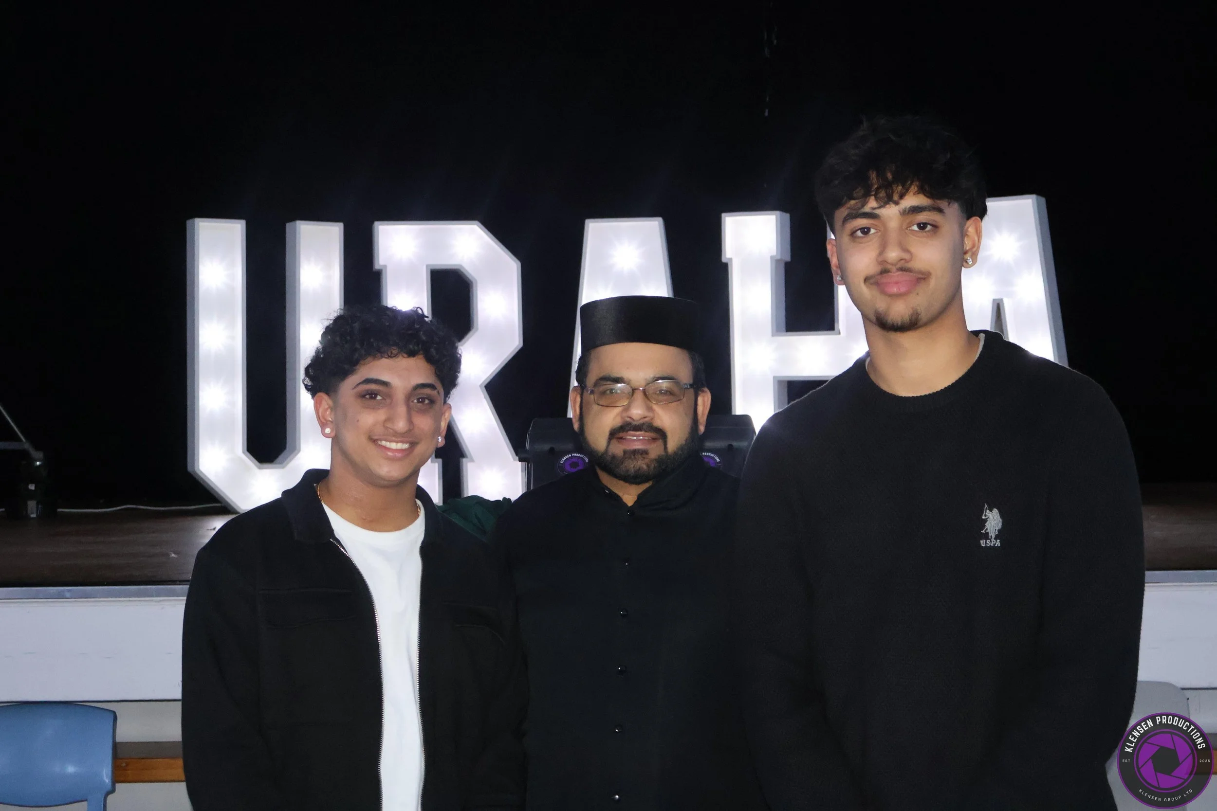 Three people standing in front of illuminated letters spelling 'UDELHIA'. The person in the center is wearing a traditional black cap and glasses, and the two people on either side are young men with short curly hair, wearing black sweaters. They are