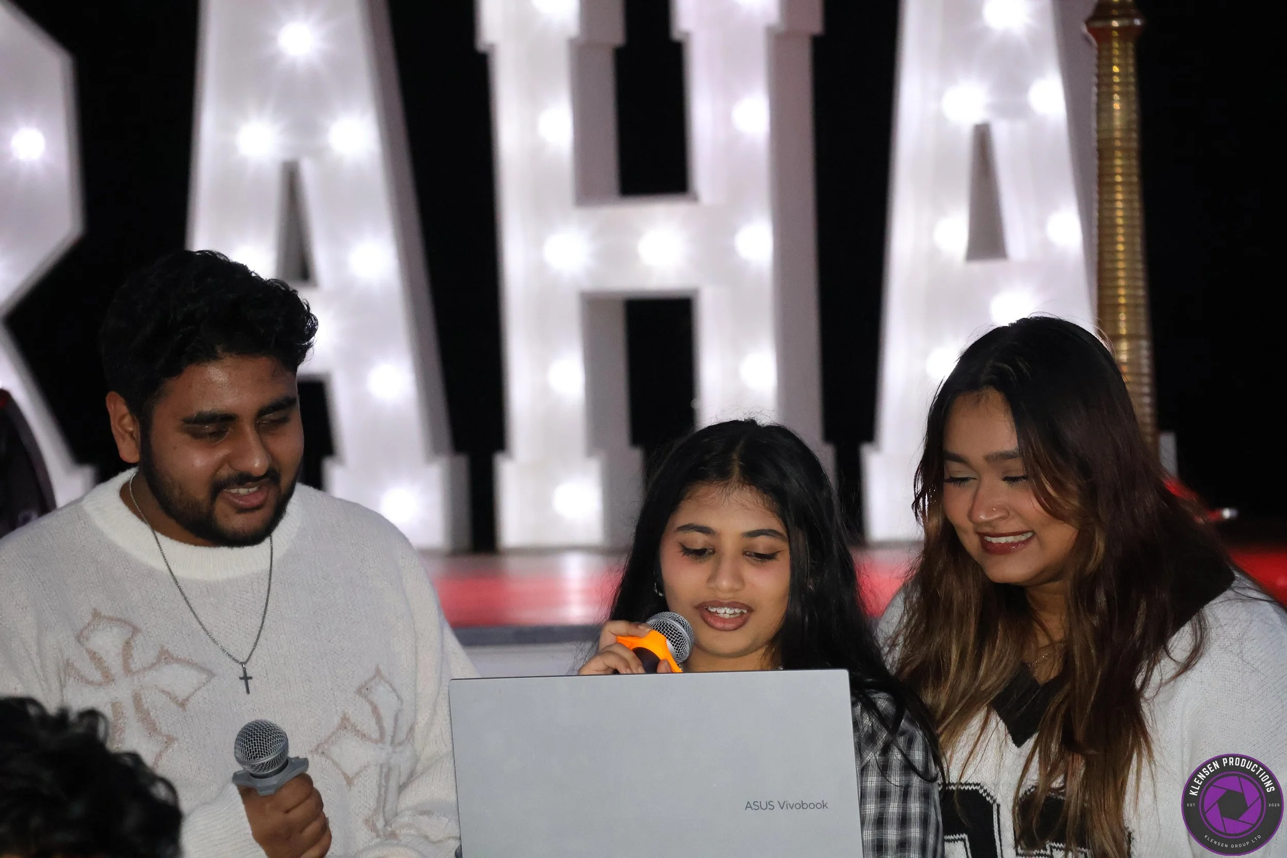 Three people, two women and a girl, are gathered around a laptop, smiling and looking at the screen. The girl is holding a microphone, and one woman is also holding a microphone. They are standing in front of a lit-up sign with large letters.