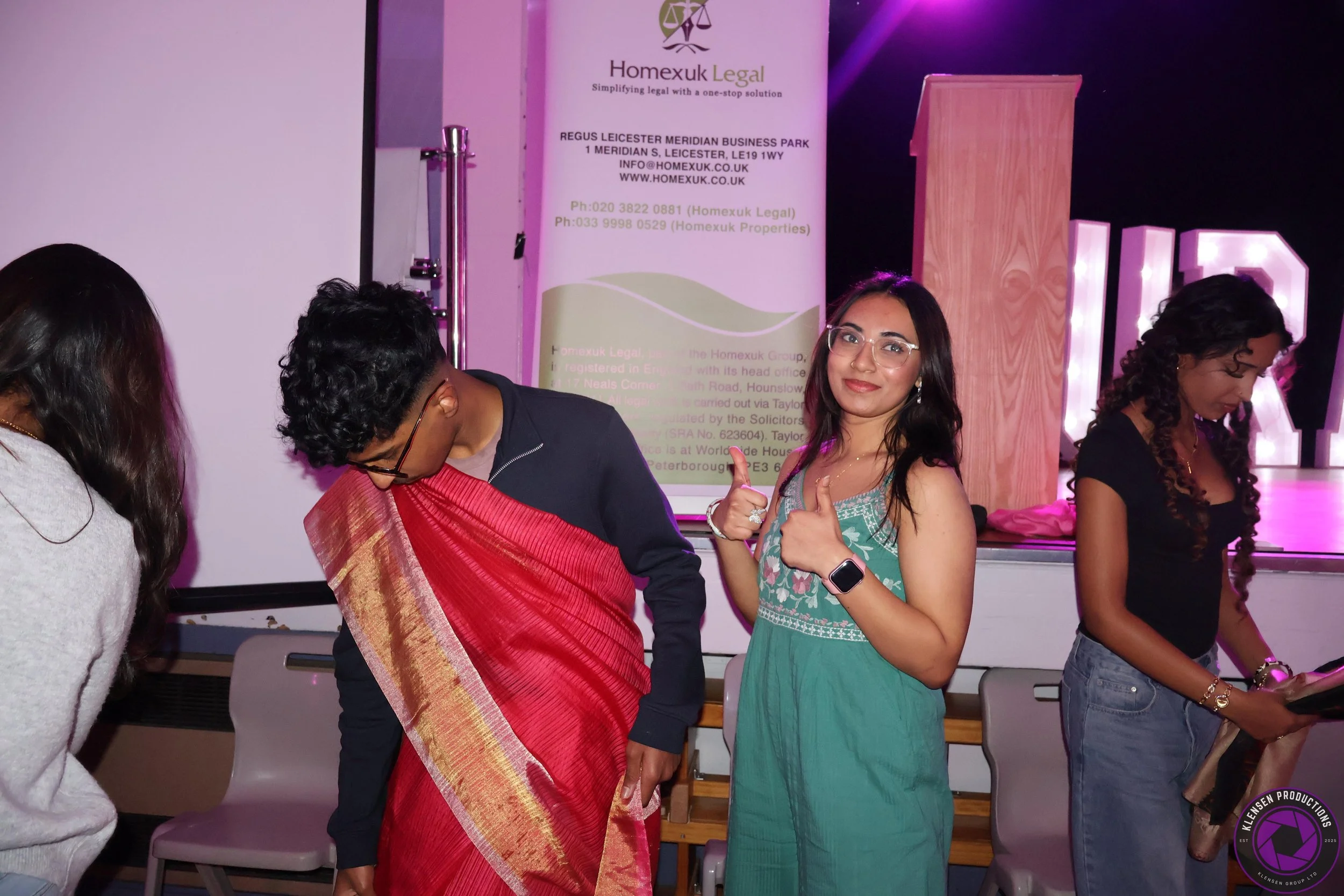 Group of women at an indoor event, with one woman in center giving a thumbs-up and looking at the camera, others looking down or away, with a banner in the background reading 'Homexuk Legal' and pink lighting.