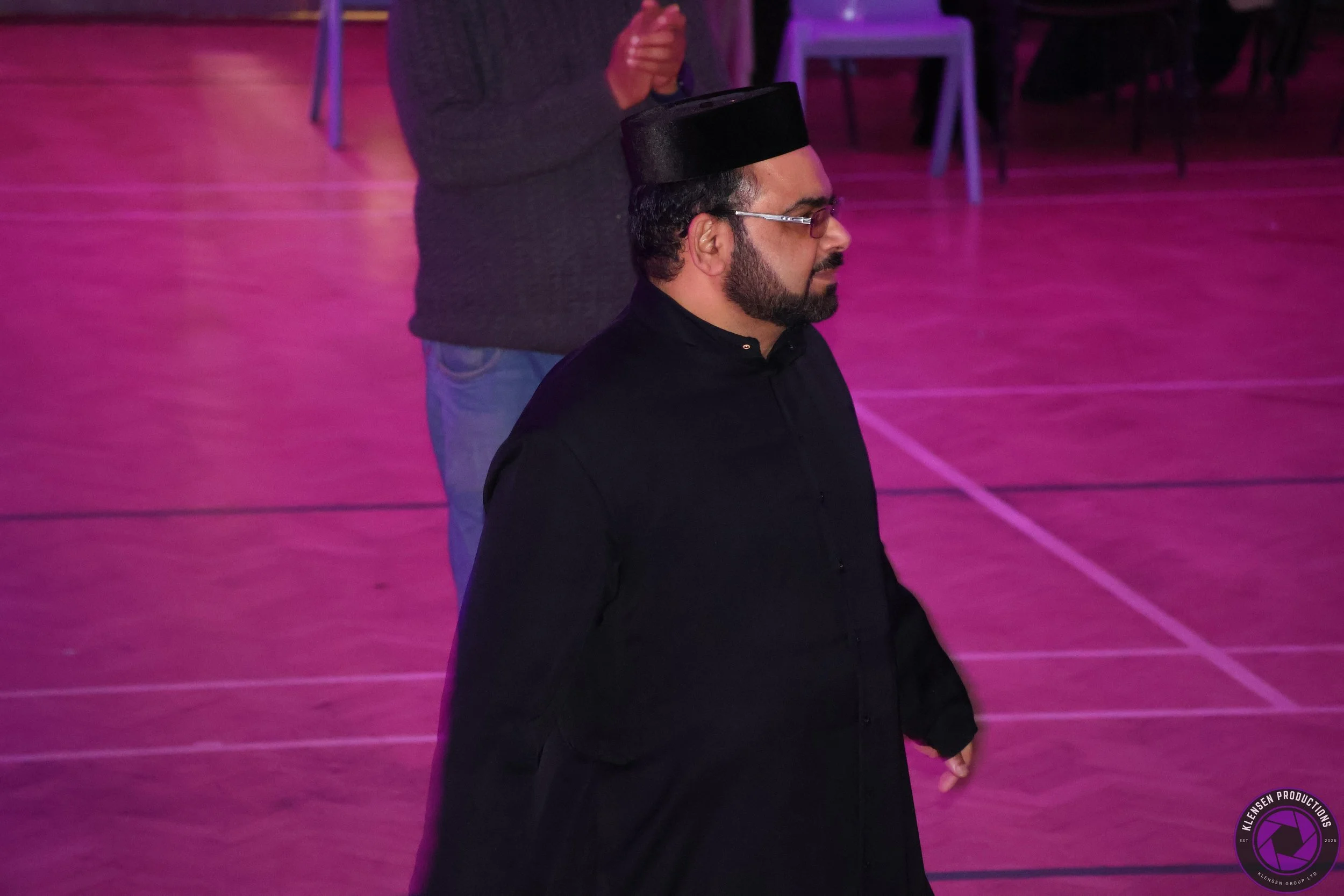 A man dressed in traditional Middle Eastern attire, wearing a black thobe and a black cap, is standing on a pink-lit floor with purple chairs in the background. He appears to be in a solemn or contemplative mood.