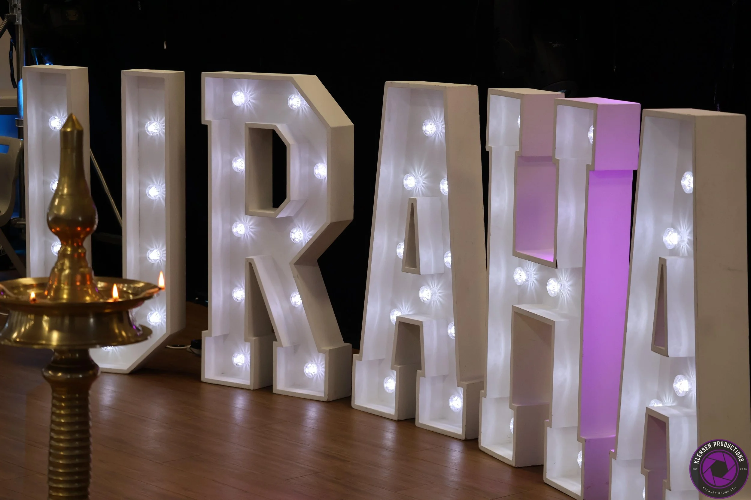 Large illuminated white marquee letters spell 'PARTY' with small light bulbs inside each letter, placed on a wooden floor against a dark background. A brass lamp with candles is partially visible in the foreground.