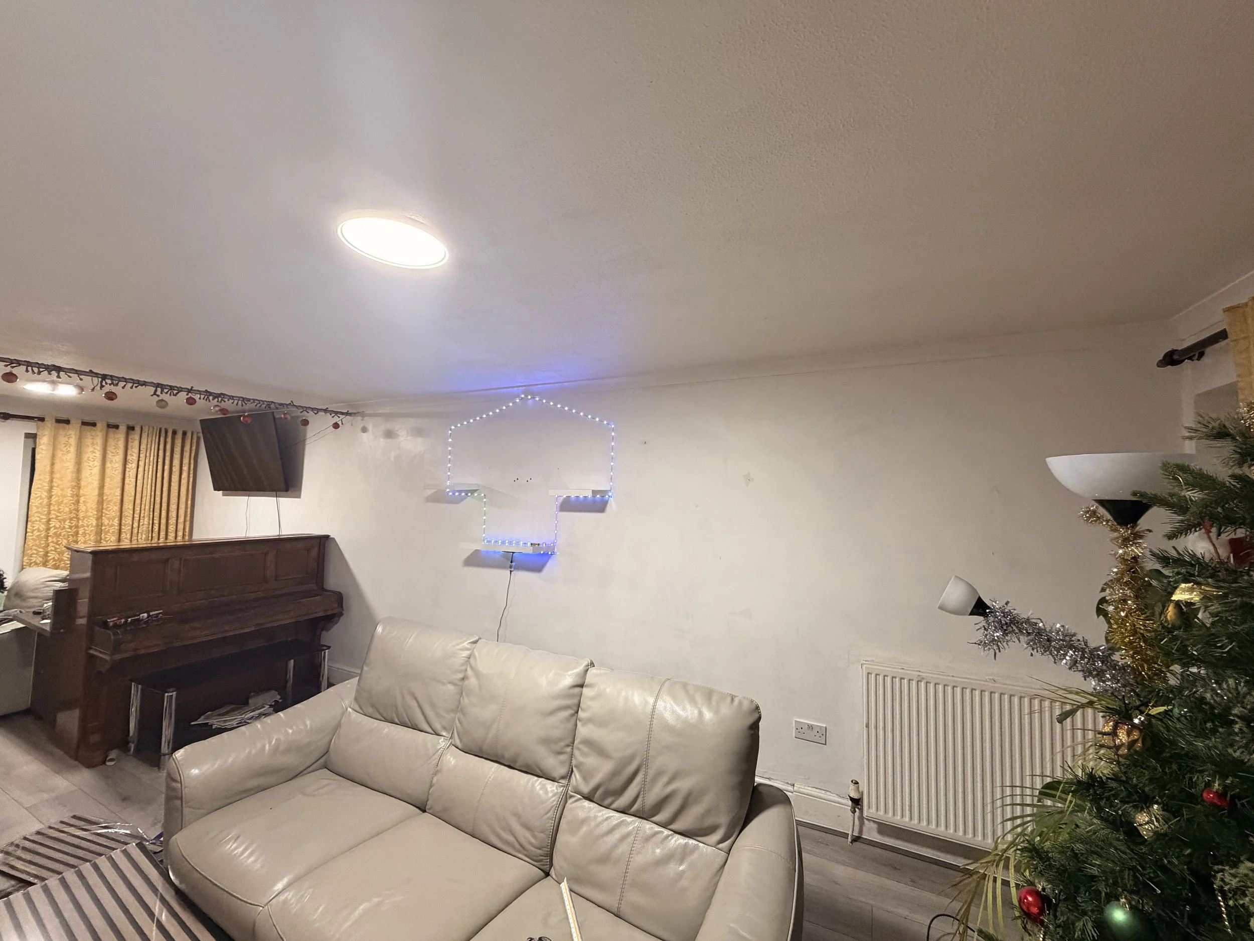 Living room decorated with Christmas lights, a Christmas tree with ornaments, a cream leather sofa, an upright piano, a wall-mounted TV, and a wall with a star-shaped light display.