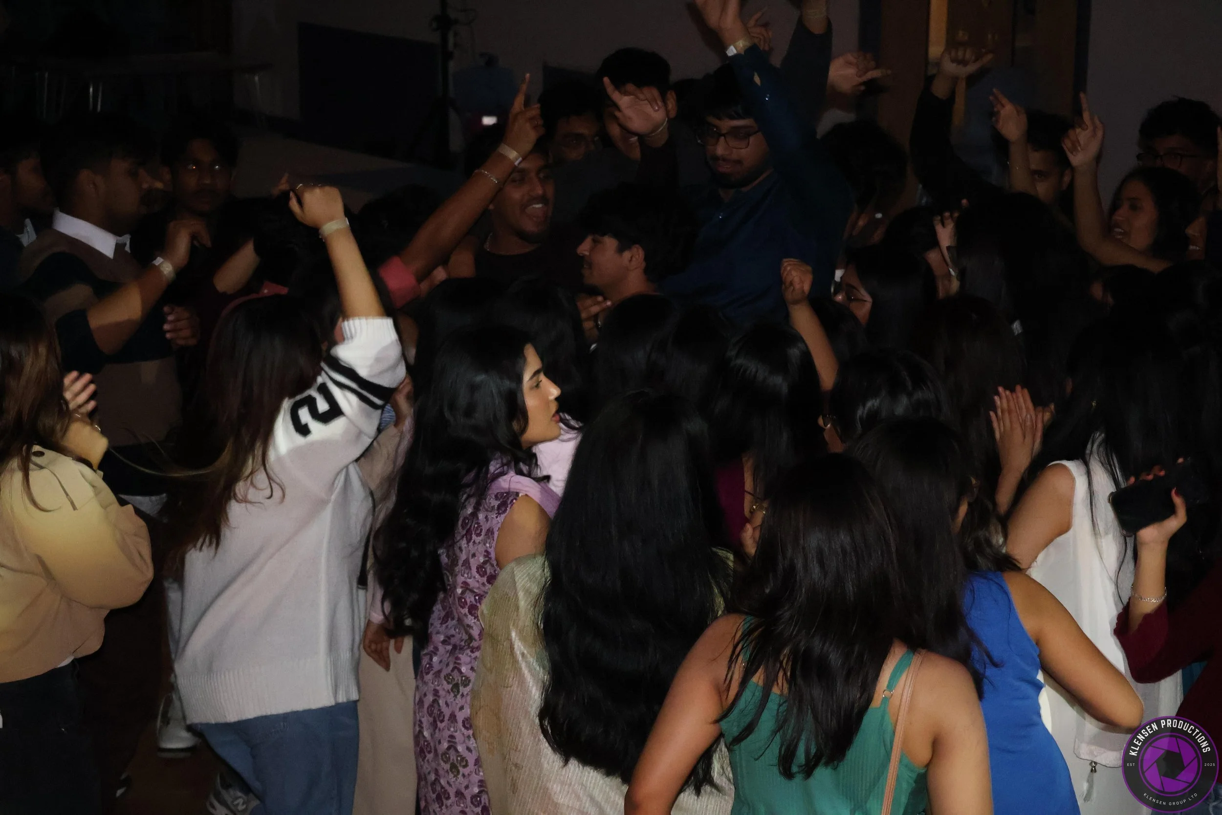 A large group of young people at a party or celebration, dancing and raising their hands in the air, with some smiling and engaging with each other.