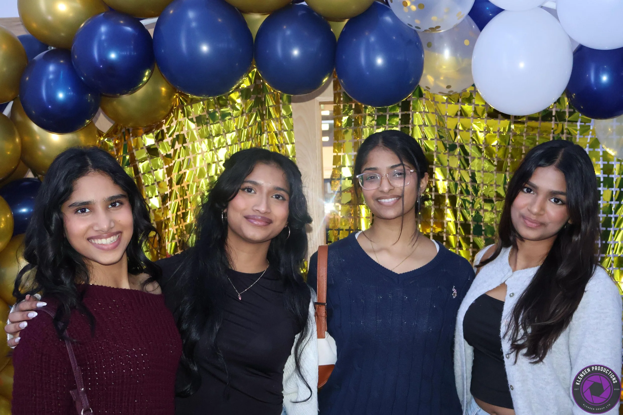 Four young women smiling at a celebration with blue, gold, and white balloons and a golden backdrop.