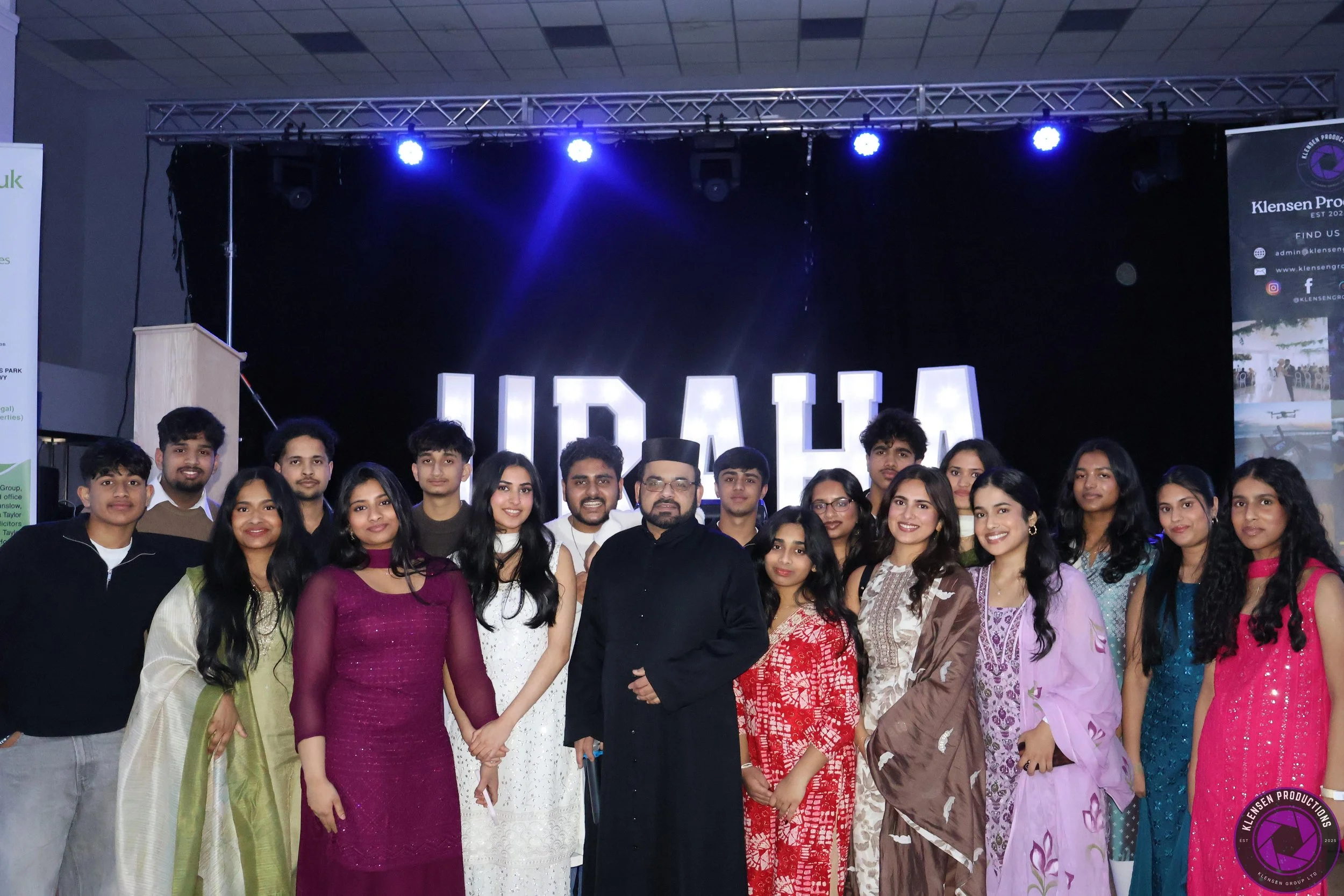 A group of young people dressed in traditional Indian attire and casual clothing posing for a photo on a stage with a black background and large illuminated letters spelling "UDAYA."