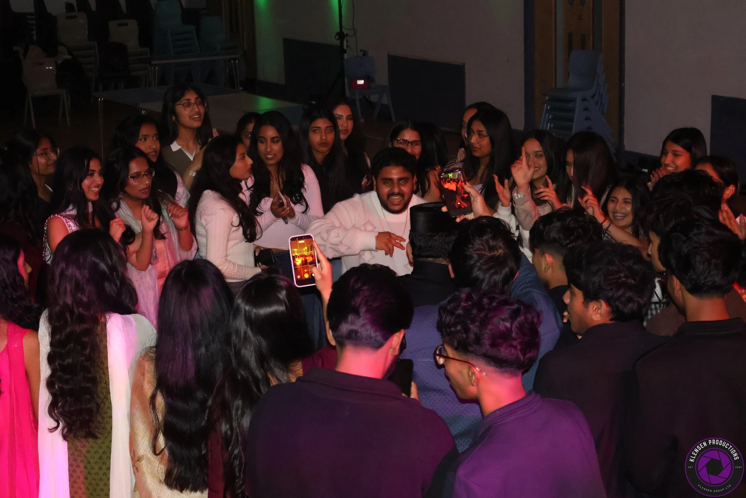 A group of young people, mostly women, gathered in a circle at an indoor event, taking pictures and selfies, one man in the center appears to be performing or speaking, with several people capturing the moment on their phones.