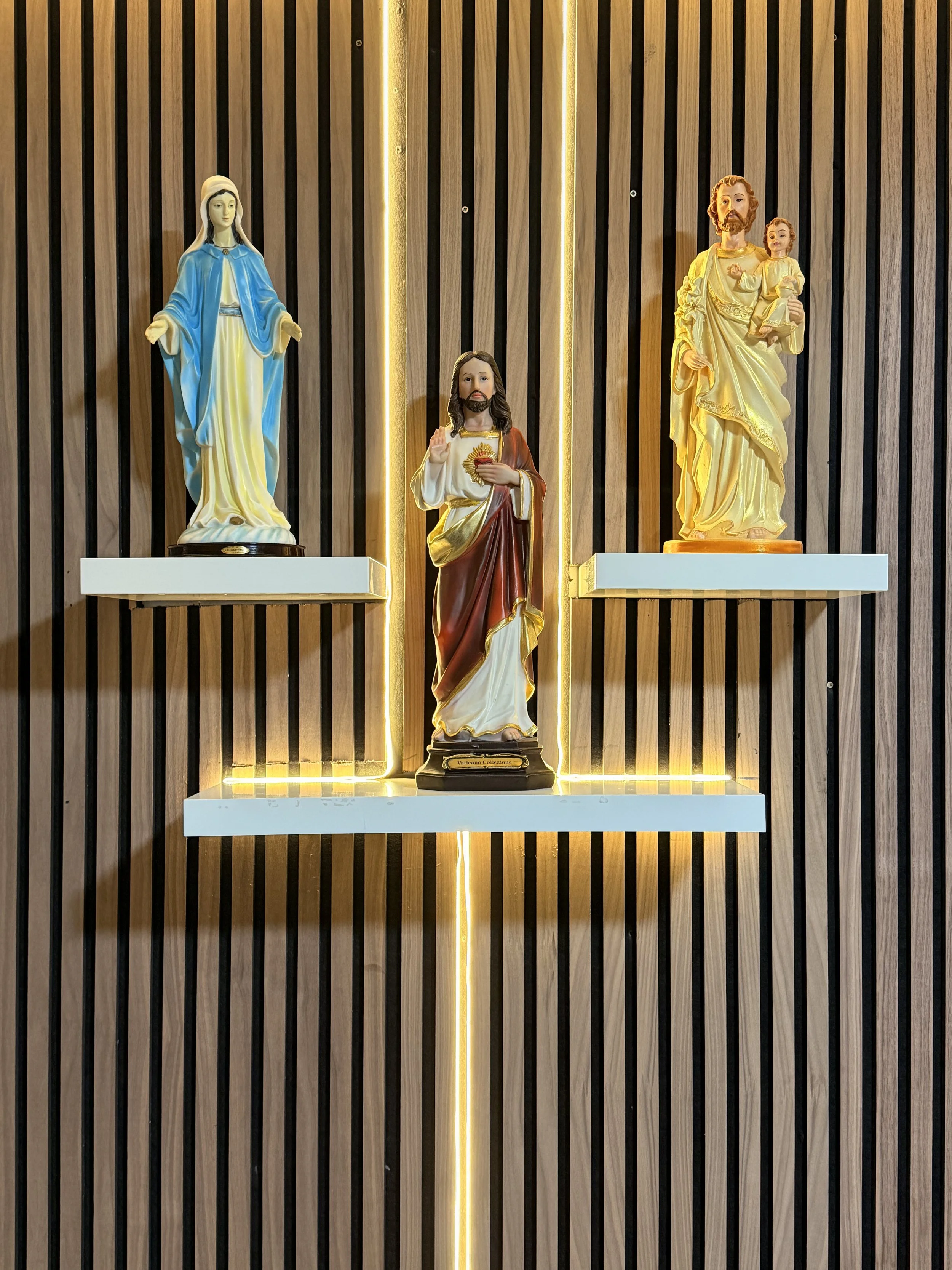 Three religious statues on wall-mounted shelves with vertical wooden slats background and LED lighting visible around them.