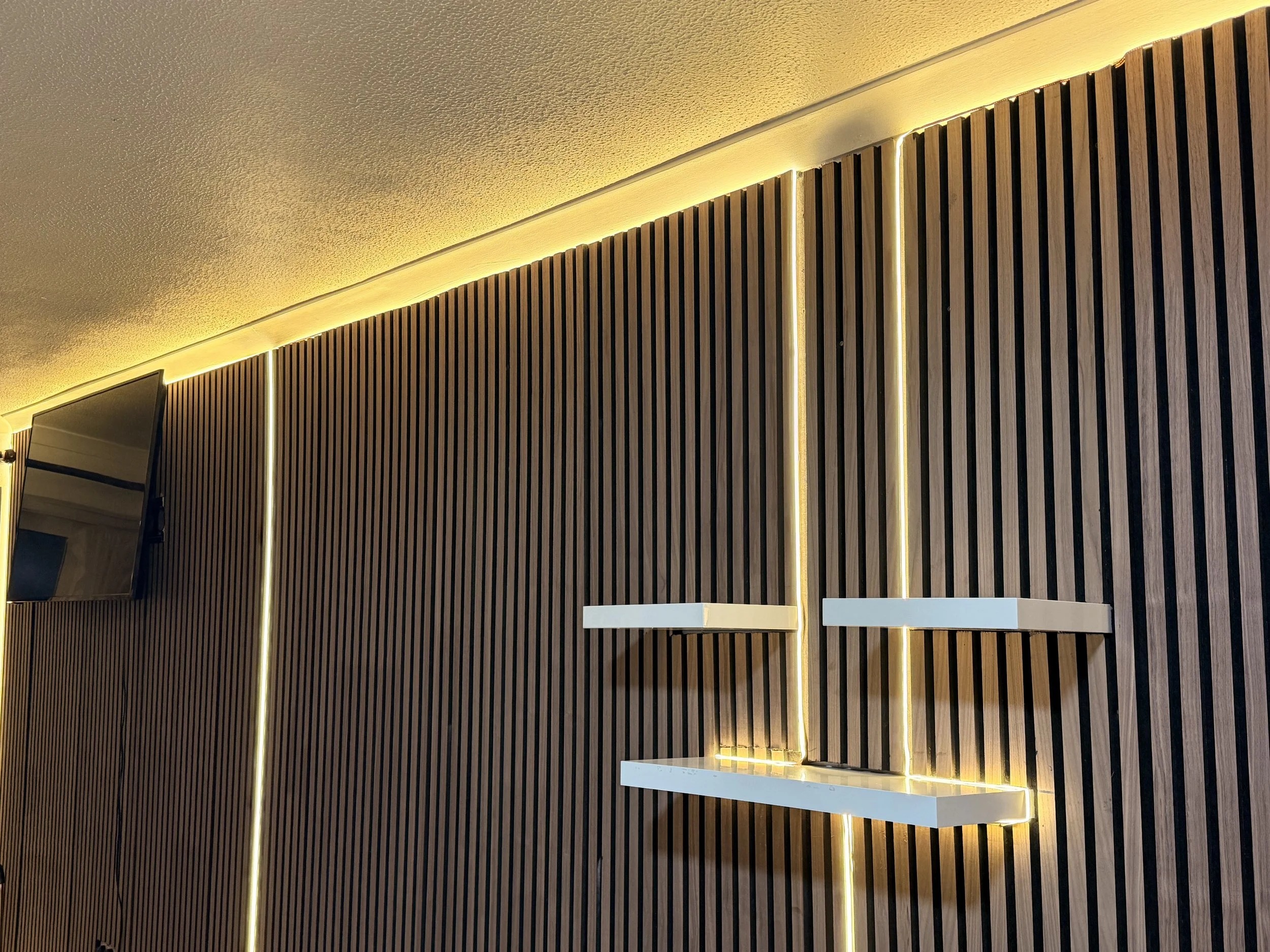 Modern interior wall with vertical wooden slats, illuminated by LED strip lighting, featuring two white floating shelves and a flat-screen TV on the left.