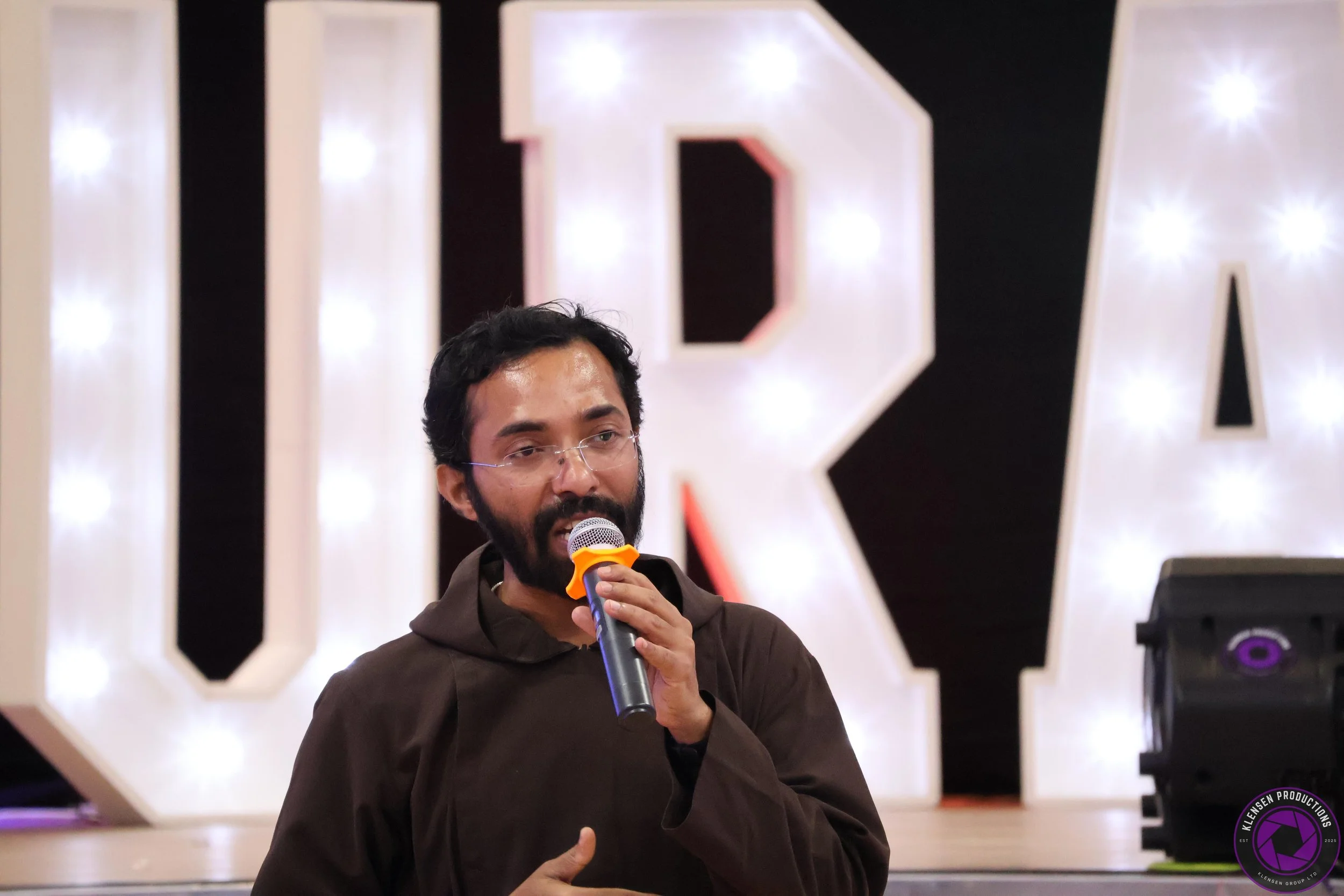 A man with glasses and a beard speaking into a microphone on stage, with large illuminated letters in the background.