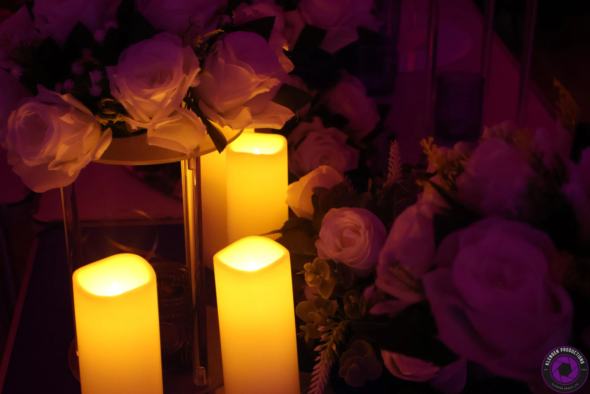 Warm yellow candle lights surrounded by soft pink roses and white floral arrangements in a dimly lit setting.