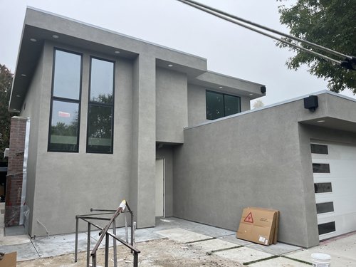 Modern gray multi-story house under construction with large windows, construction materials, and tools visible in the foreground.