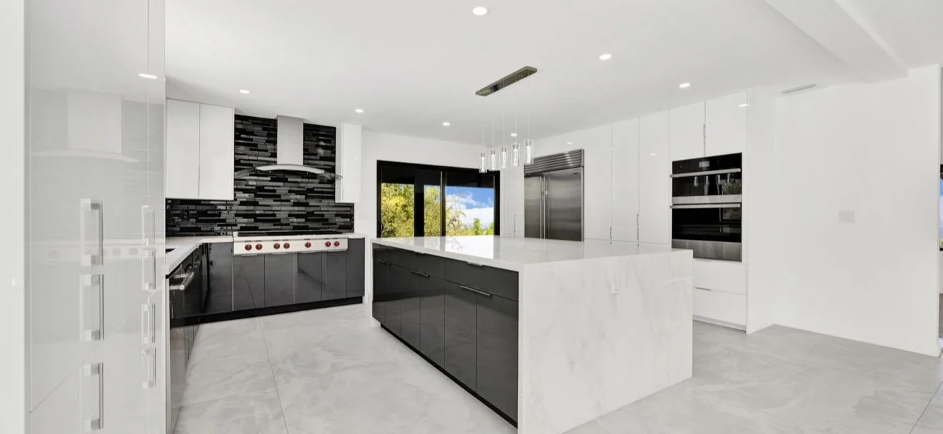 Finished kitchen with white upper cabinets, dark lower cabinets, a light stone island, patterned backsplash and a large window.