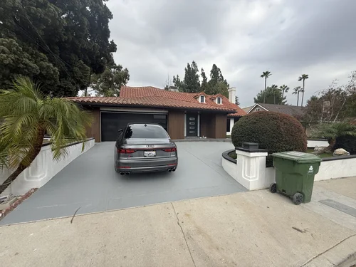 Residential house with a red-tile roof, a black garage door, and a black car parked on the driveway. Surrounding greenery includes large trees and trimmed bushes. The sky is overcast and cloudy.