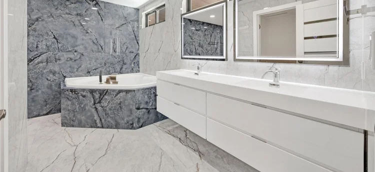 Modern bathroom with a white double sink vanity, large mirrors, and a corner bathtub with dark grey marble walls.