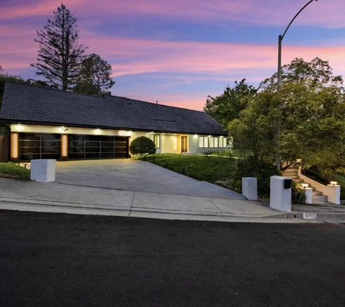 Modern house with illuminated entrance, driveway, landscaped yard, stairs leading to the side, and a sunset sky with pink and purple hues.