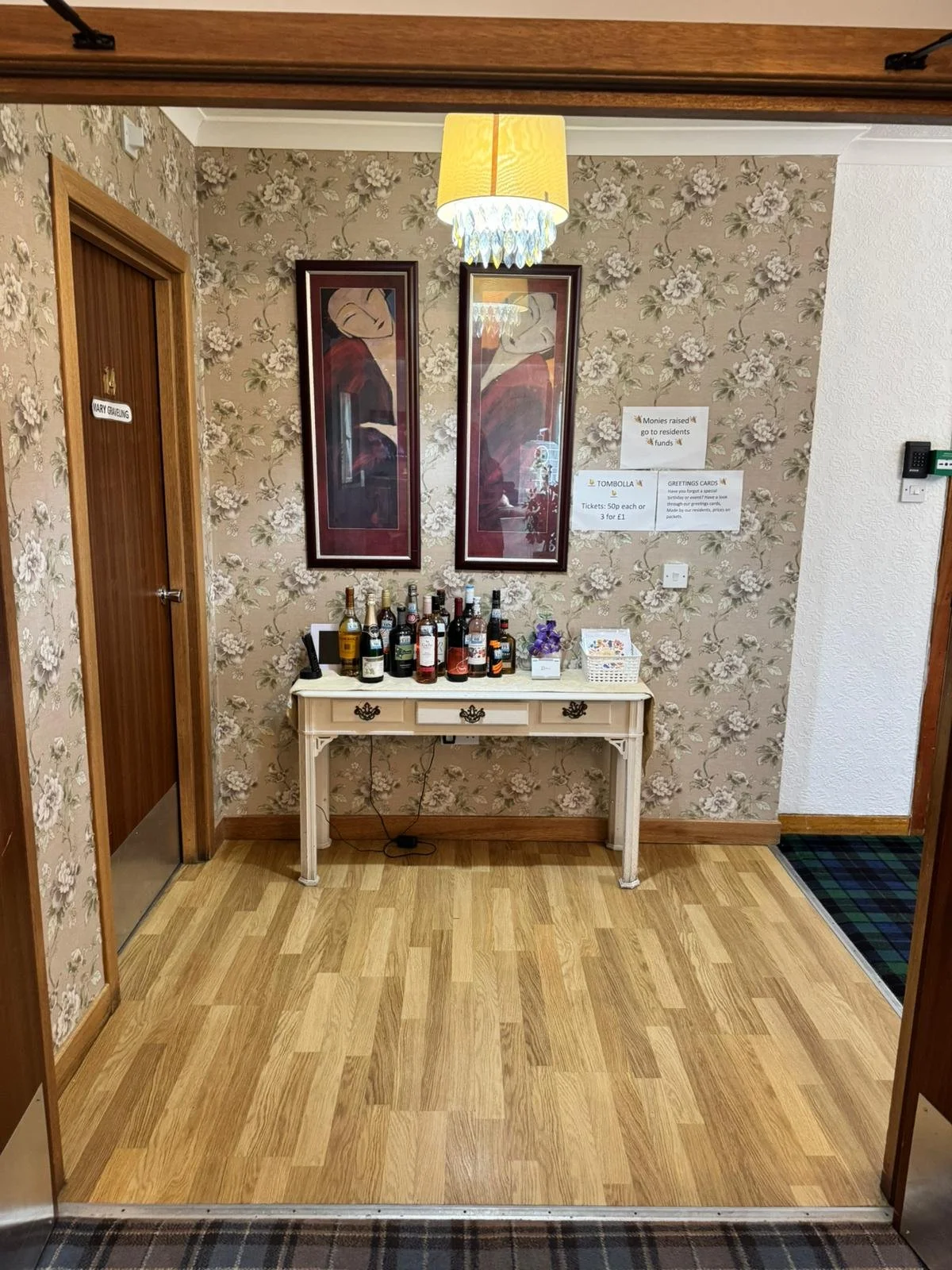 A small table with several bottles of alcohol, decorated with a small plant and some candies, stands in front of a floral wallpapered wall with two framed paintings. Signs on the wall advertise selling greeting cards and tombola tickets, and a bright