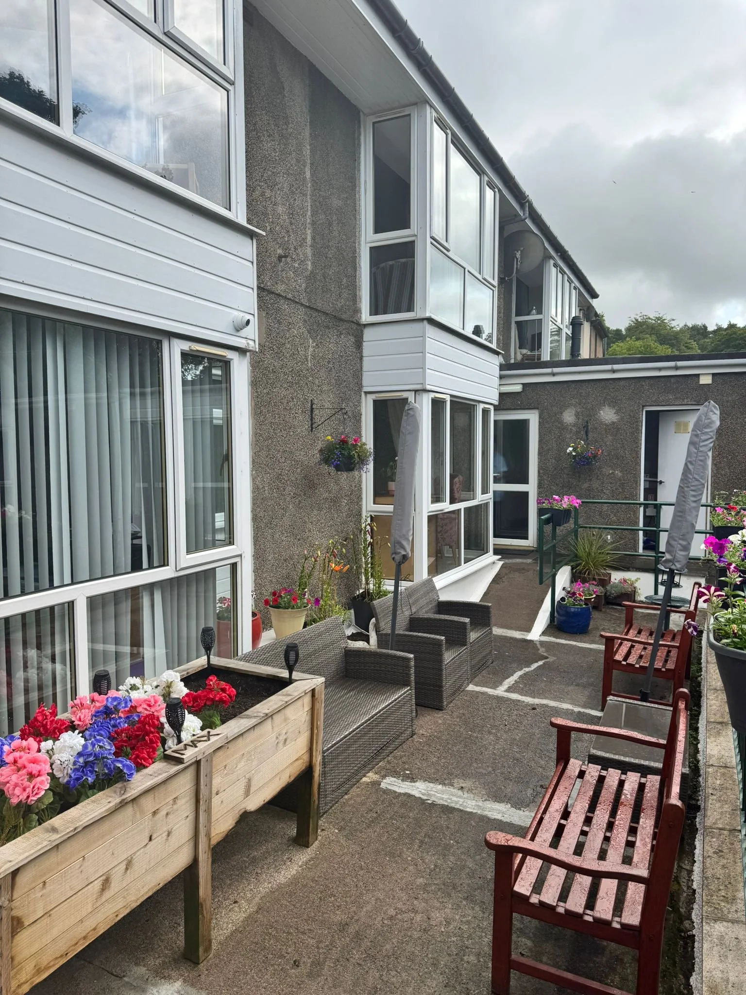 Outdoor patio area with colorful flower planters, wicker chairs, wooden benches, and closed patio umbrellas, adjacent to an apartment building with large windows and balconies.