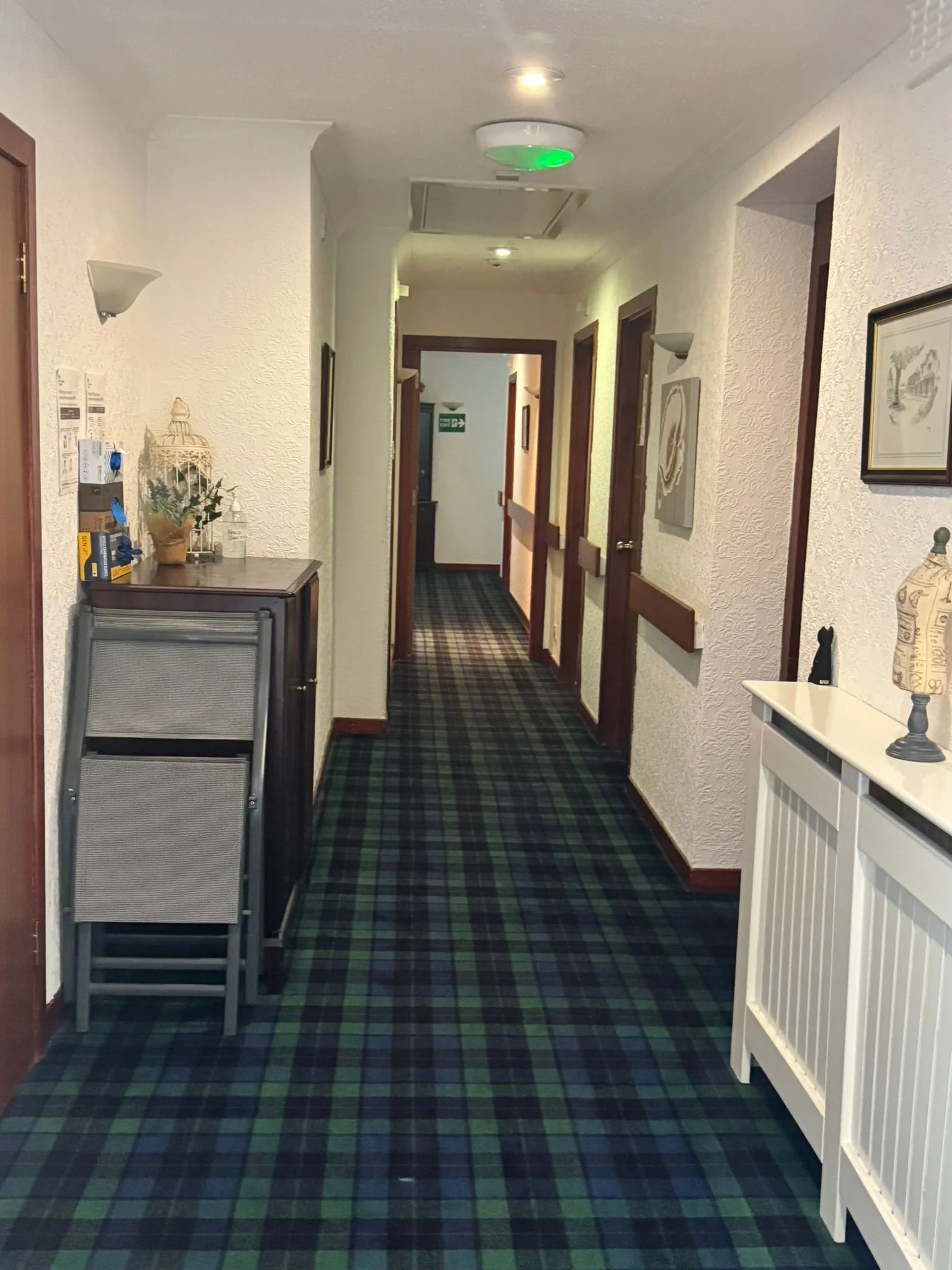 Long hotel corridor with patterned green and blue carpet, white walls, wooden door frames, wall-mounted lights, and decorative items on small tables.