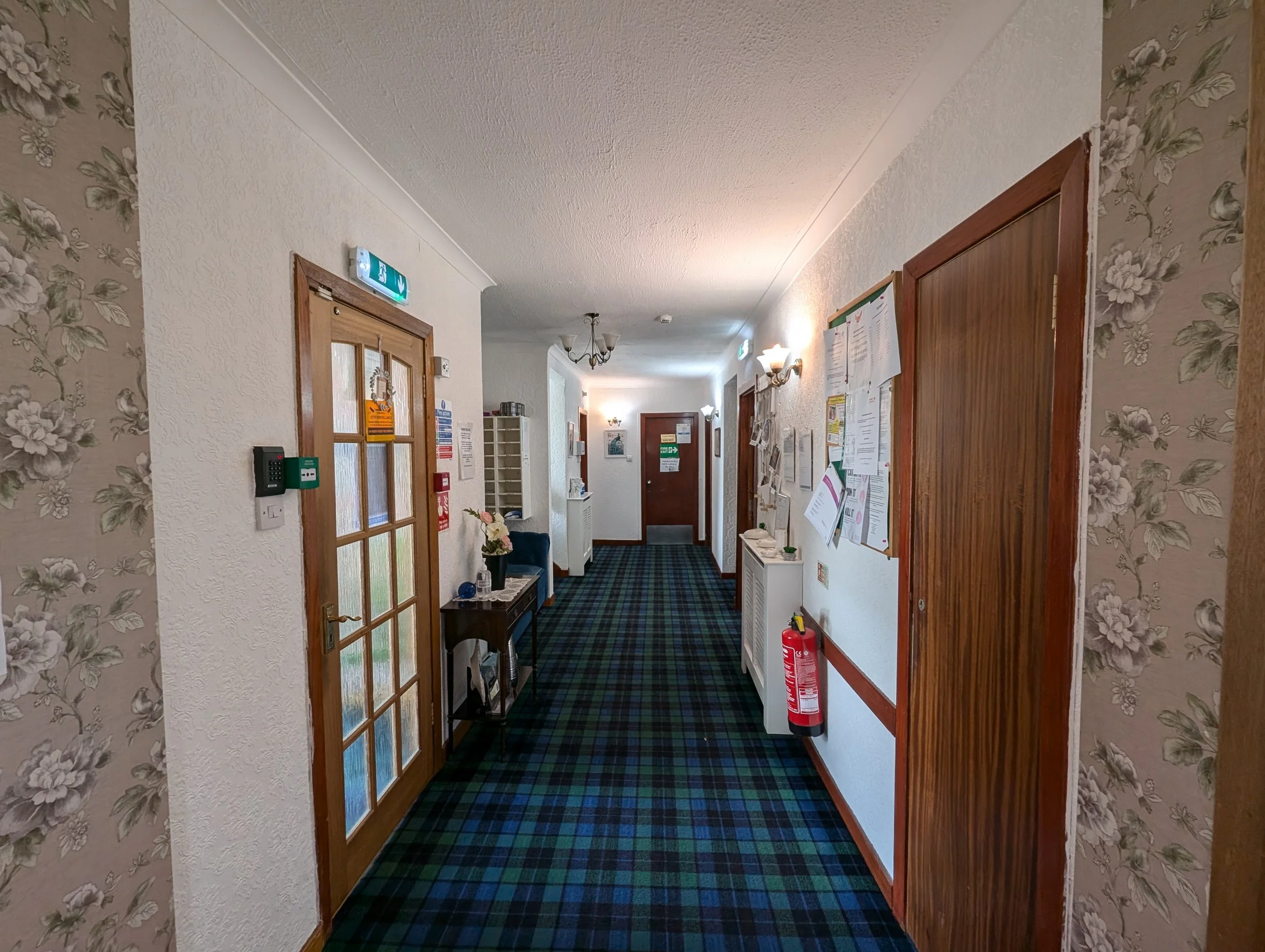 Hallway in a cozy inn or hotel with floral wallpaper, carpeted floor in a tartan pattern, wall-mounted lights, and bulletin boards.