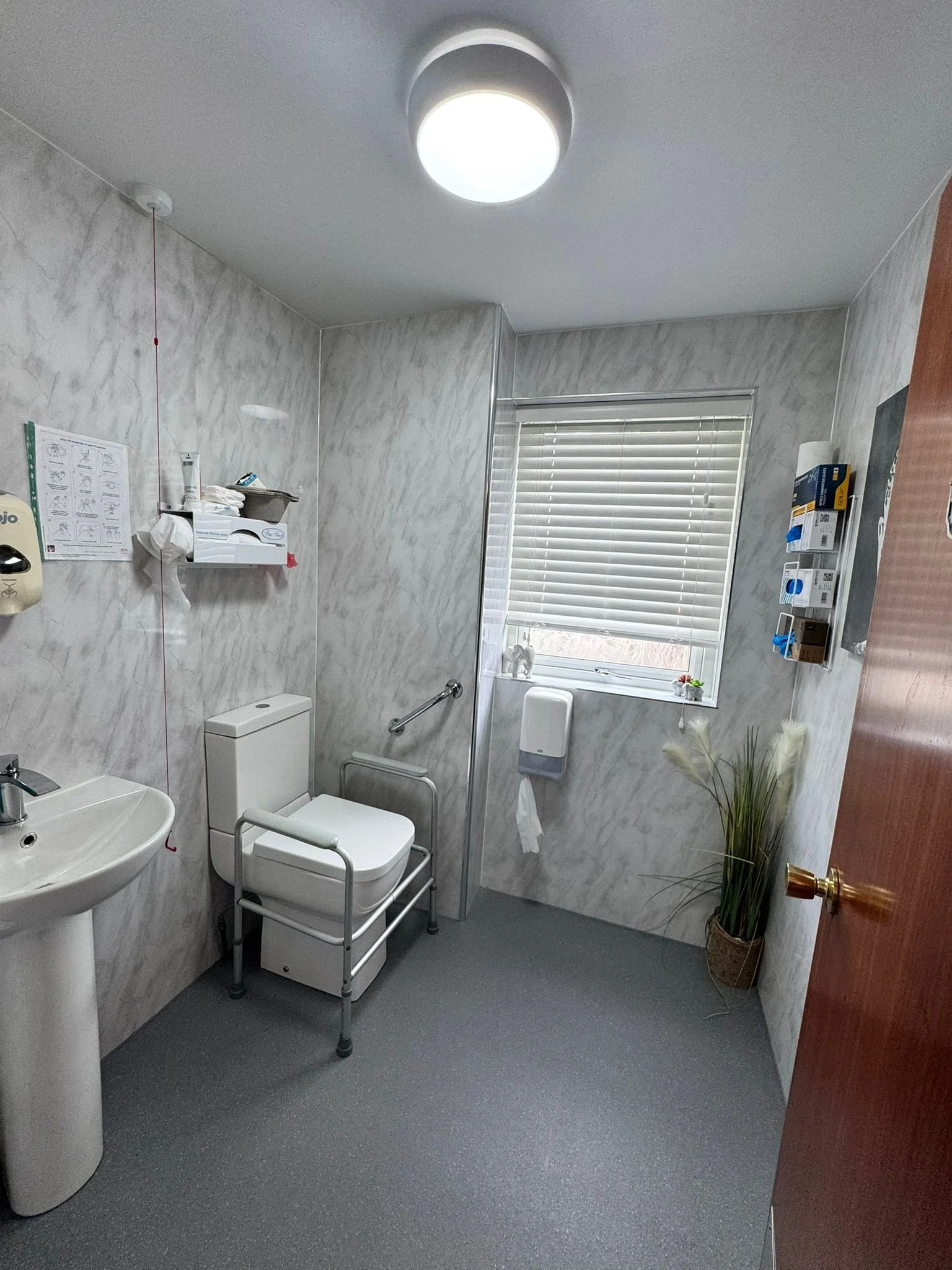 A hospital or care facility bathroom with a white toilet, a sink, a window with blinds, medical supplies on the wall, a potted plant, and accessible grab bars.