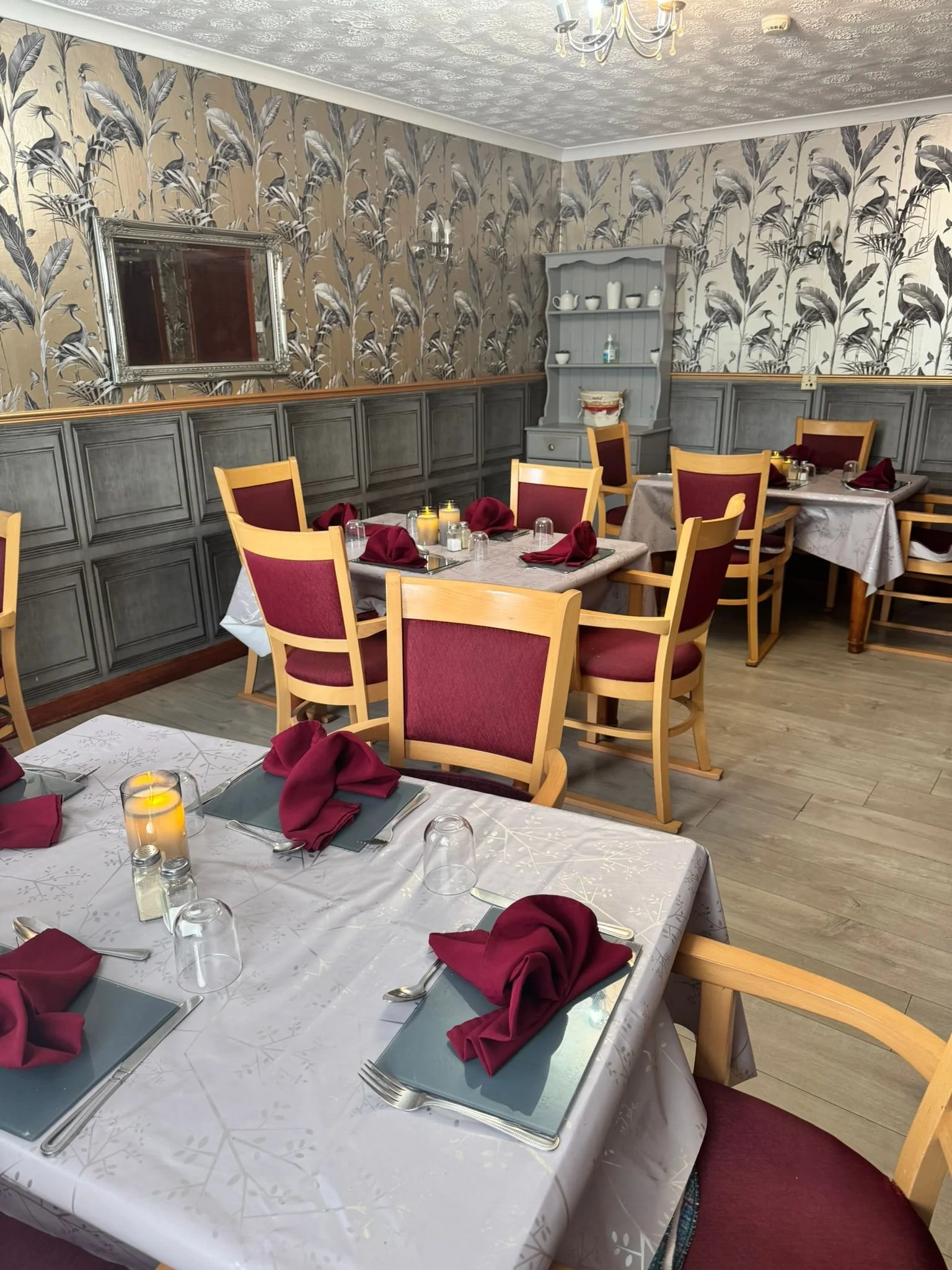 Dining room with tables set for a meal, featuring burgundy napkins, candles, and place settings, with patterned wallpaper and wooden chairs with burgundy cushions.