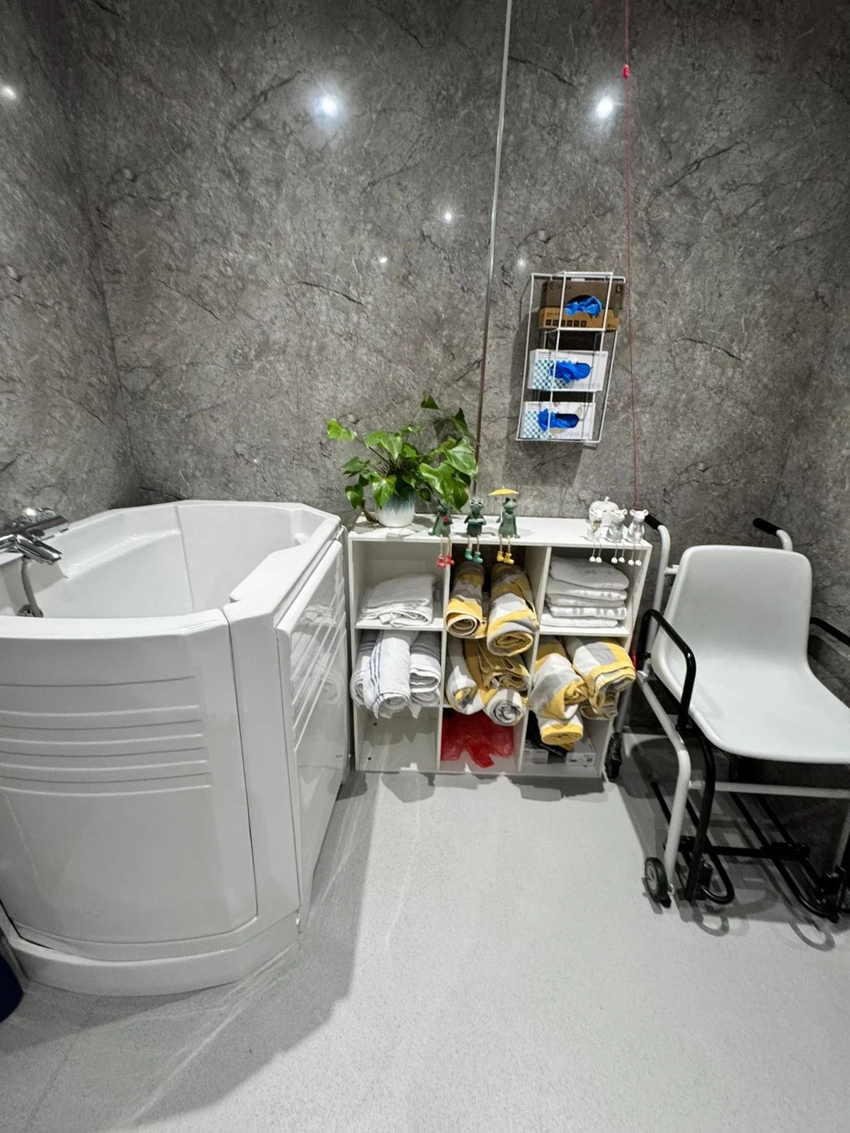 A room with a medical or spa setting, featuring a white soaking tub, a collection of rolled yellow and gray towels, a white shelf with folded white towels, a potted green plant, and a white chair with black metal armrests. A gray marble wall with cei